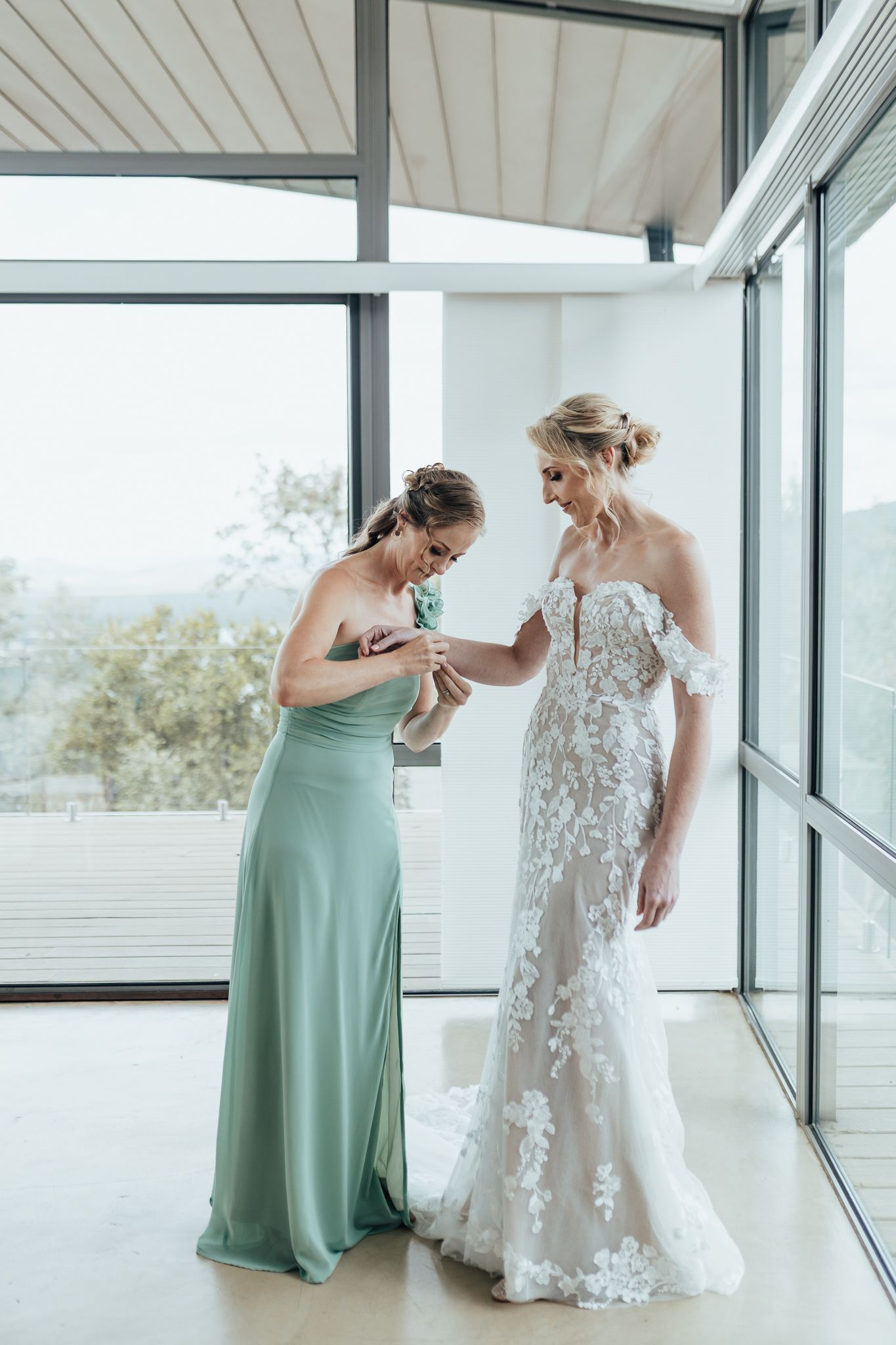 Wedding dress moment shared between bridesmaid in mint green and bride in lace gown by sunlit windows.