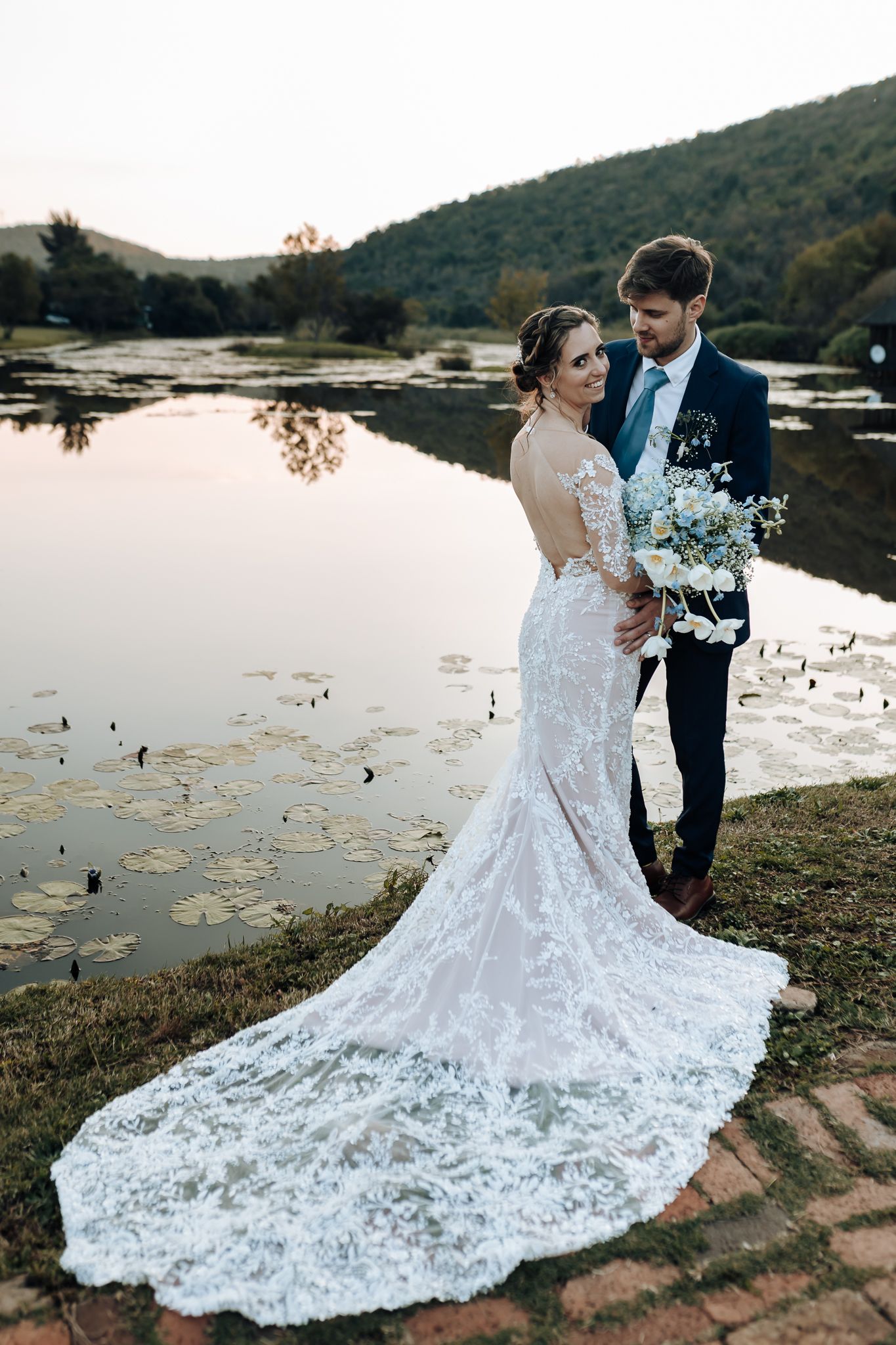 Newlyweds pose together by a tranquil lake, with the bride wearing a stunning lace gown with long train.