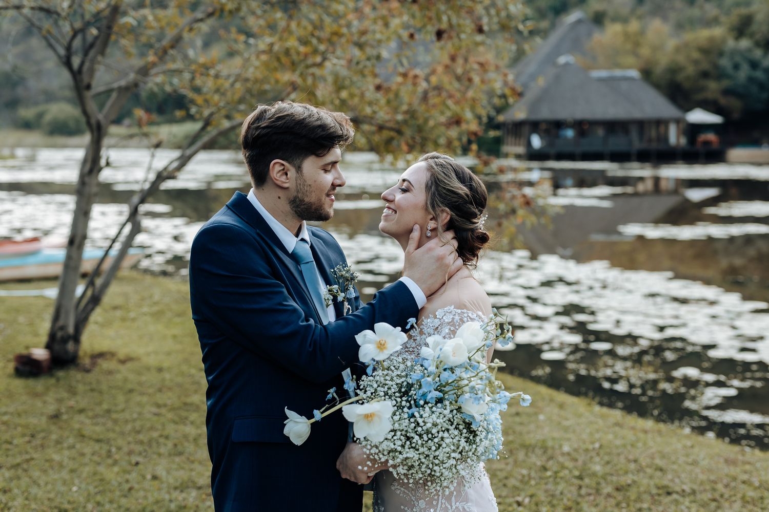 Wedding couple embraces by a lake with autumn trees and a rustic cabin in the background.