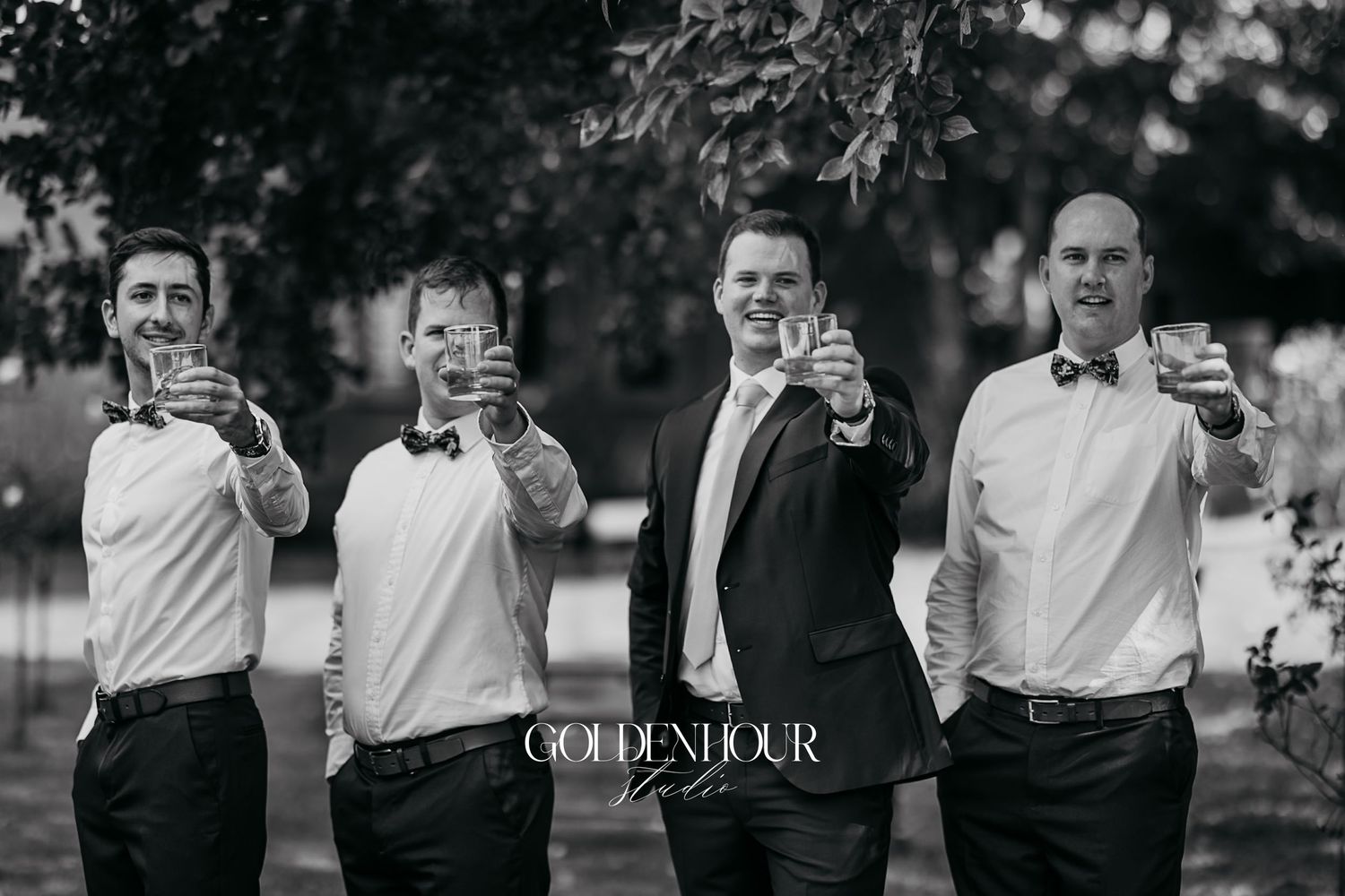 Groom and groomsmen in bow ties raise glasses in a celebratory outdoor toast in black and white.