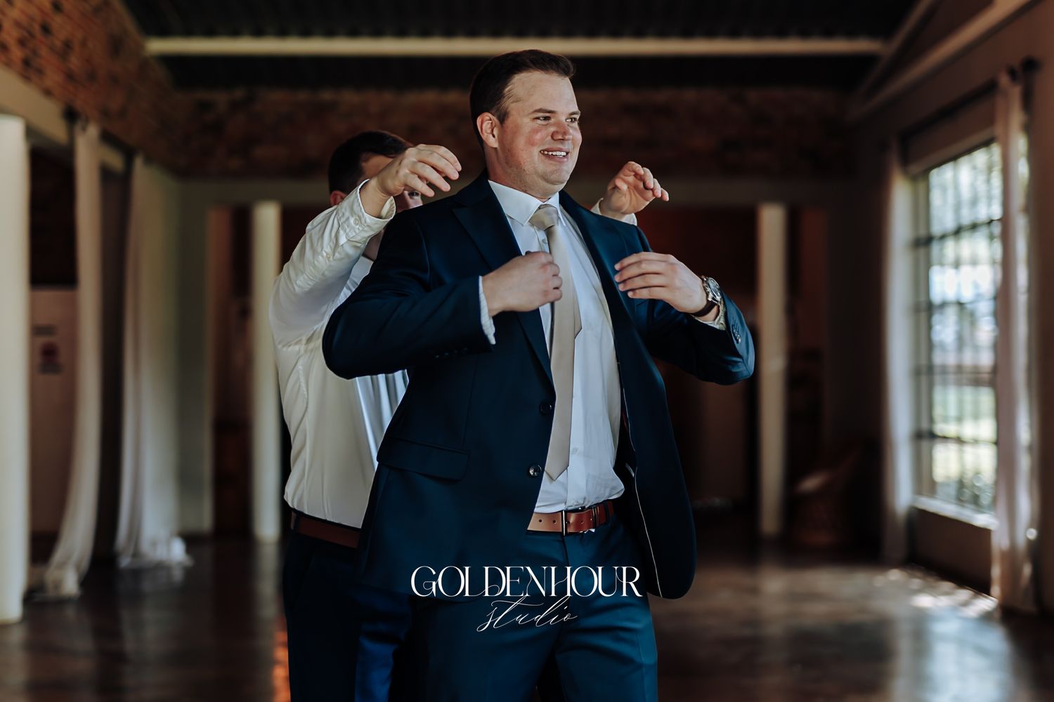 Groomsman helping the groom adjust his navy suit jacket inside a rustic brick wedding venue.