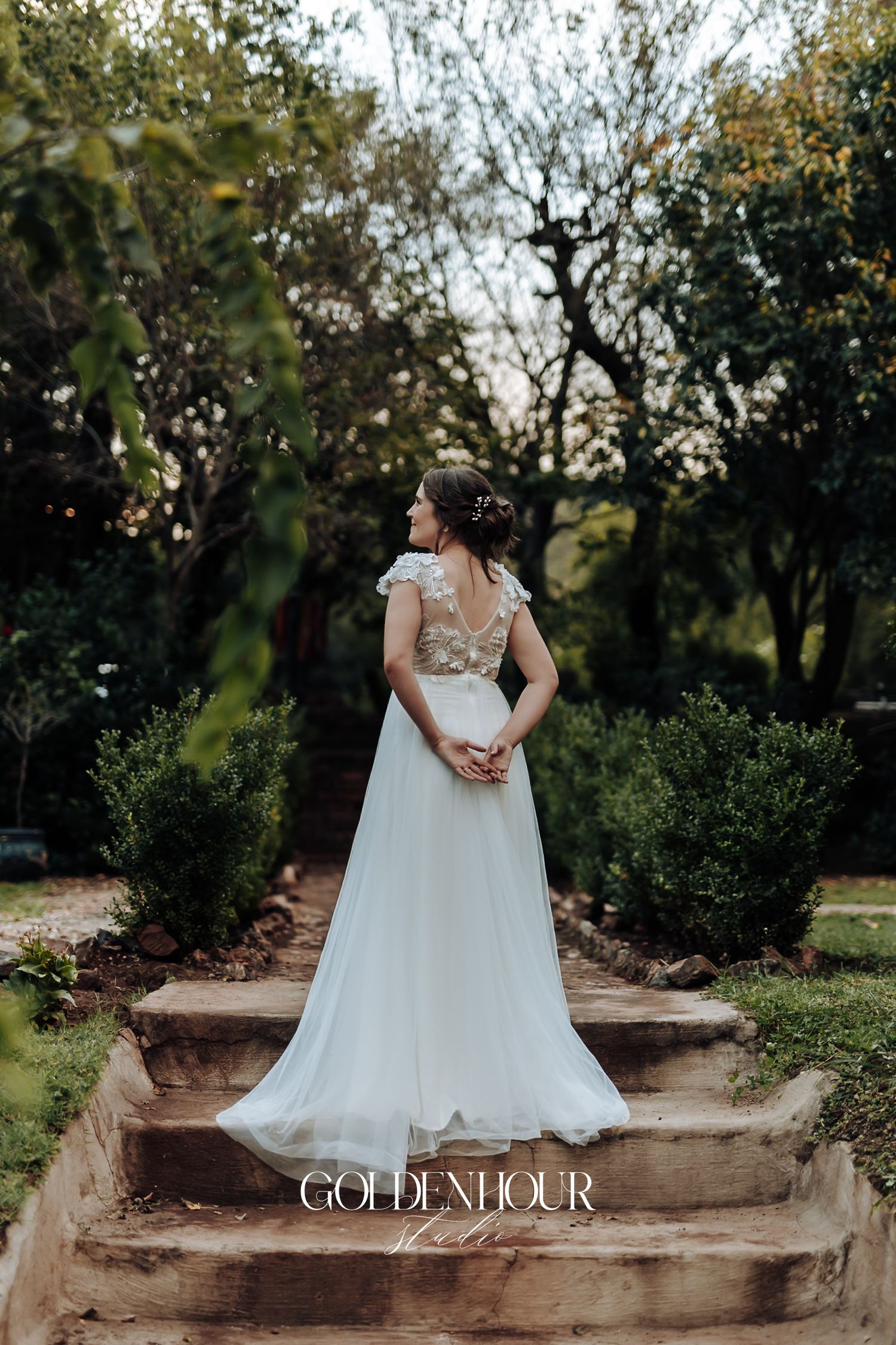 Bride in elegant two-tone wedding gown with gold embroidered bodice stands on stone garden steps surrounded by greenery.
