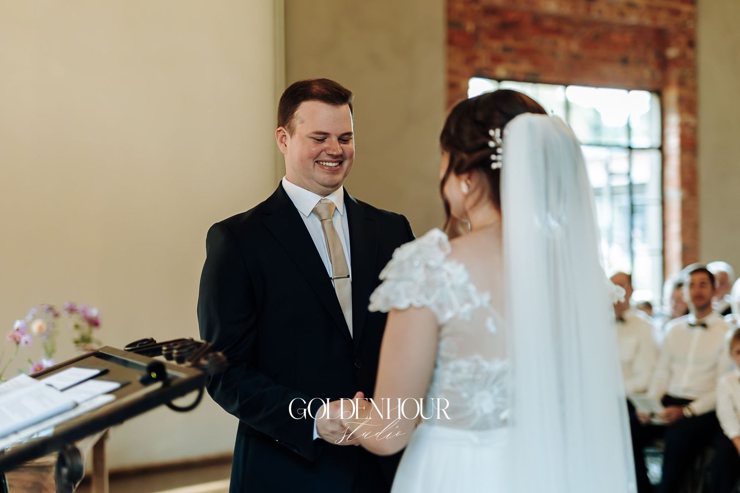 Groom in navy suit smiling at bride during indoor wedding ceremony at decorated altar.