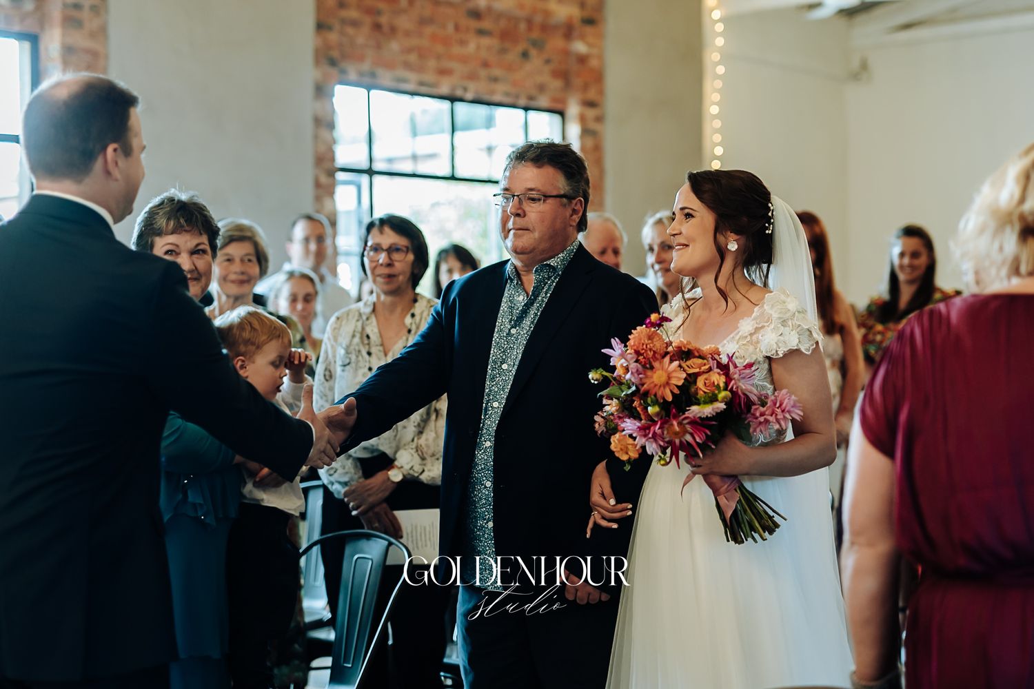 Bride holding vibrant bouquet walks down aisle with father toward groom at indoor wedding.