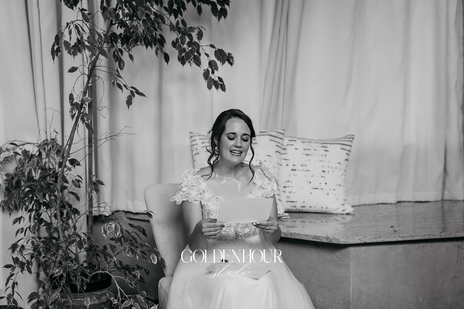 Bride in white lace wedding gown sits reading a card in a softly lit bridal suite.