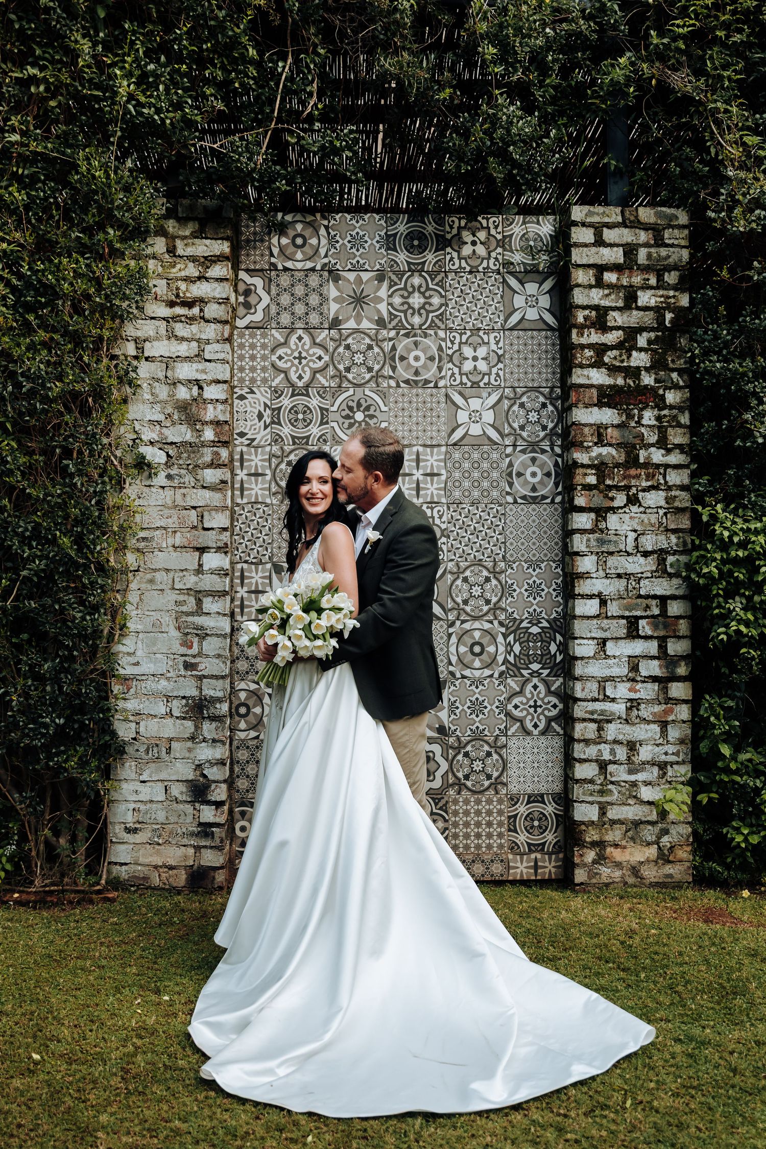 Bride and groom embrace before decorative tiled wall backdrop surrounded by lush greenery, bride holding white bouquet.