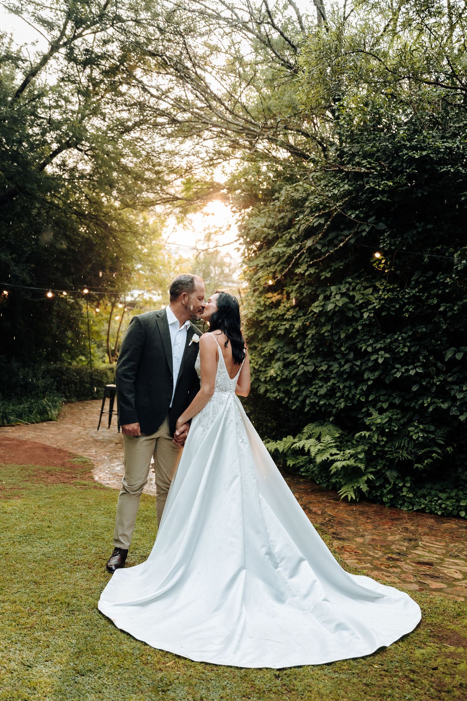 Bride and groom share a romantic kiss in a lush garden setting, with the bride's elegant white gown trailing on the ground.