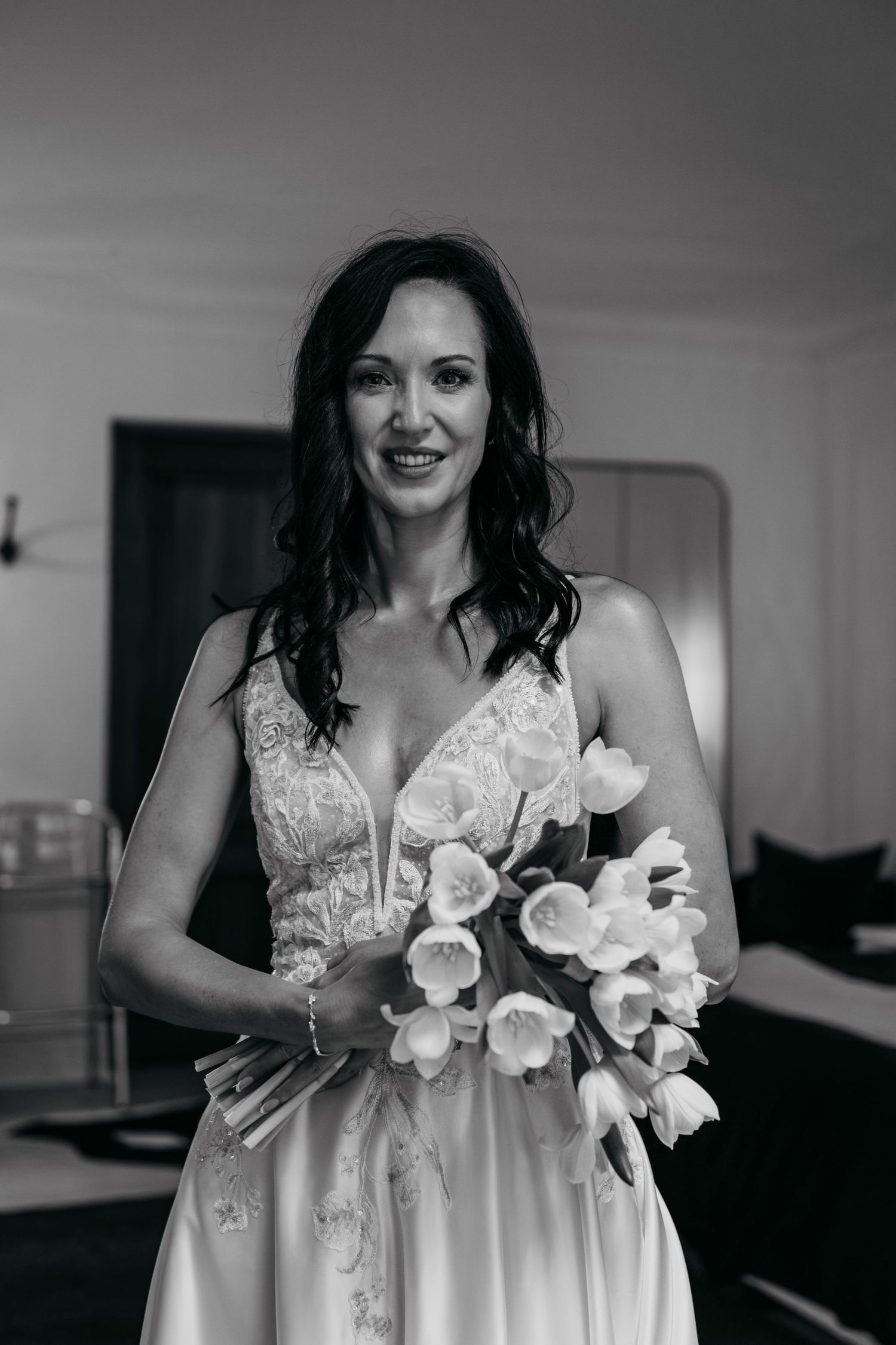 Black and white photo of a smiling bride in a lace gown holding a bouquet of tulips indoors.