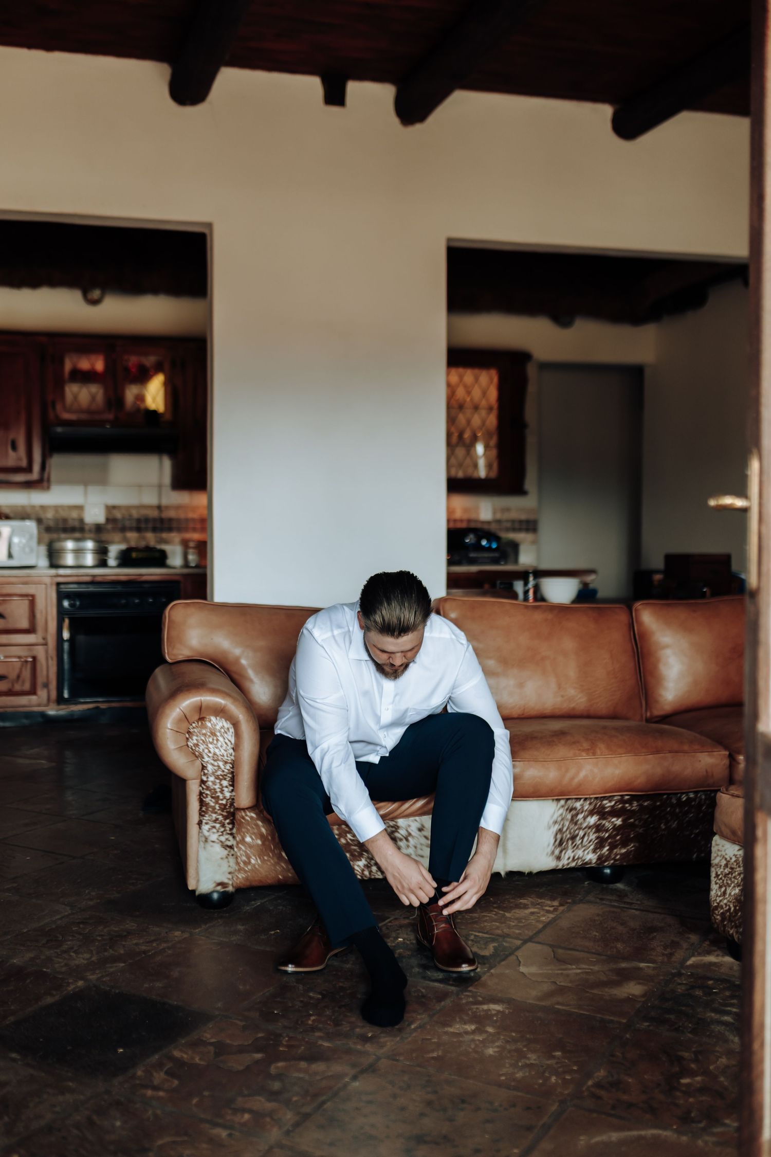A figure in white shirt and dark pants sits alone in contemplation on the edge of a couch in a dimly lit room.