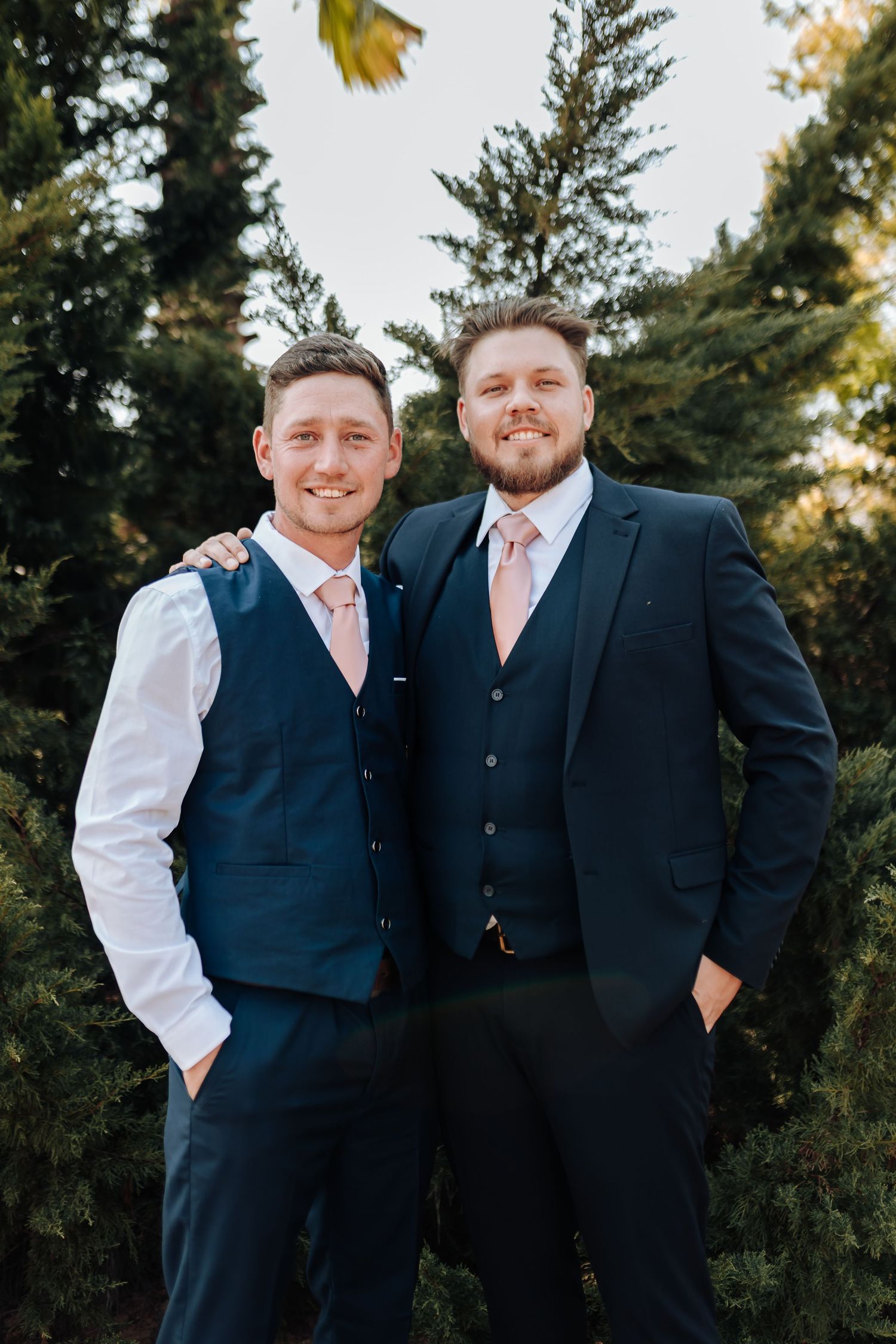 Two men in formal navy suits with pink ties pose together in front of evergreen trees on a sunny day.