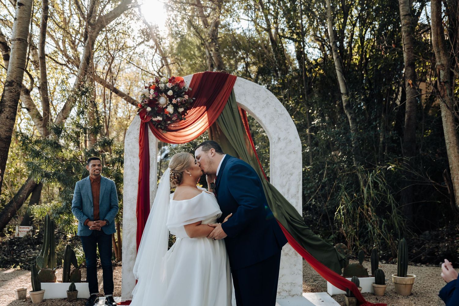 Bride and groom share first kiss under floral arch with red draping in outdoor woodland wedding ceremony.