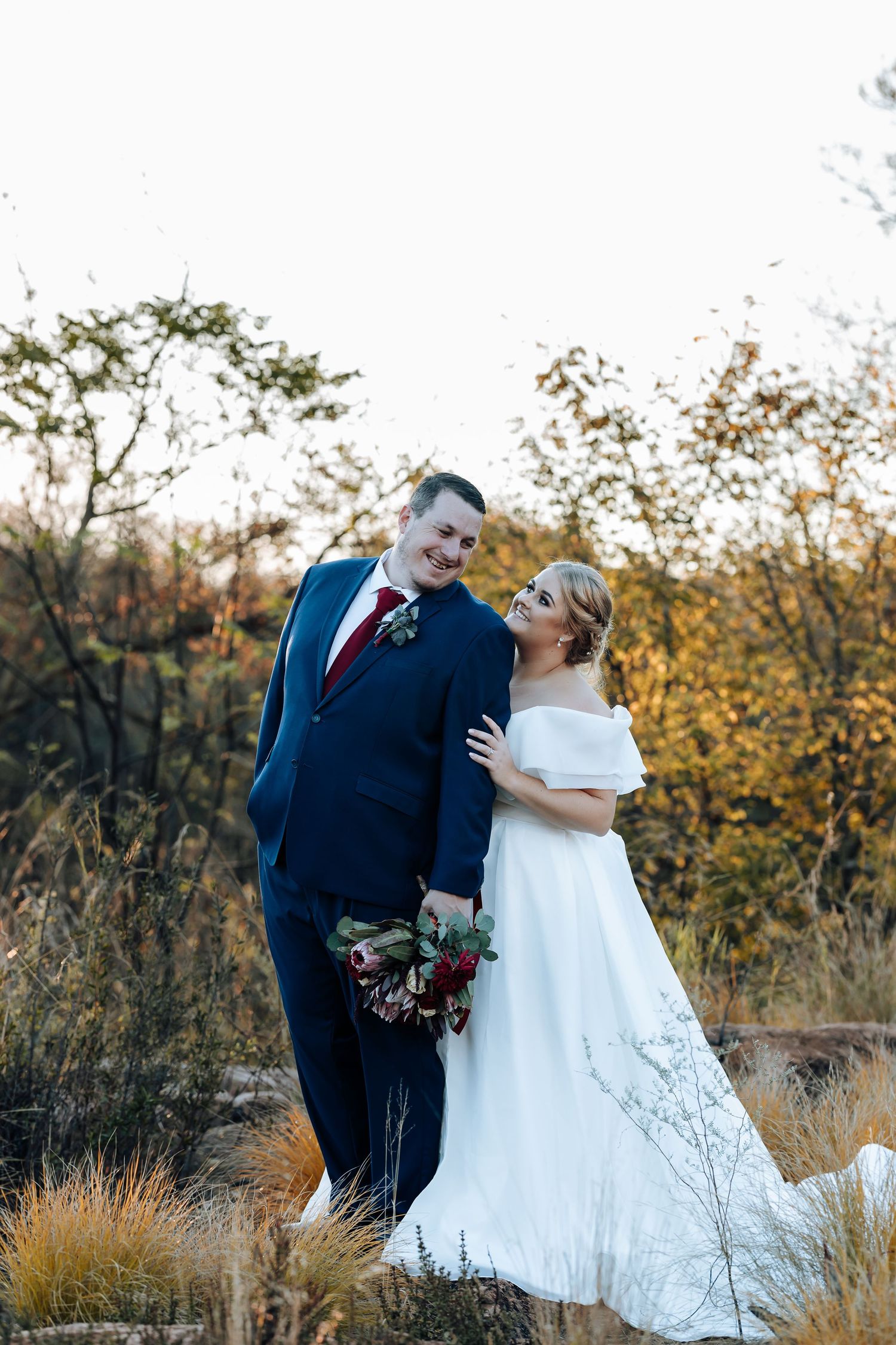 Bride and groom pose outdoors in autumn setting, she wears white off-shoulder gown, he wears navy suit with red tie.