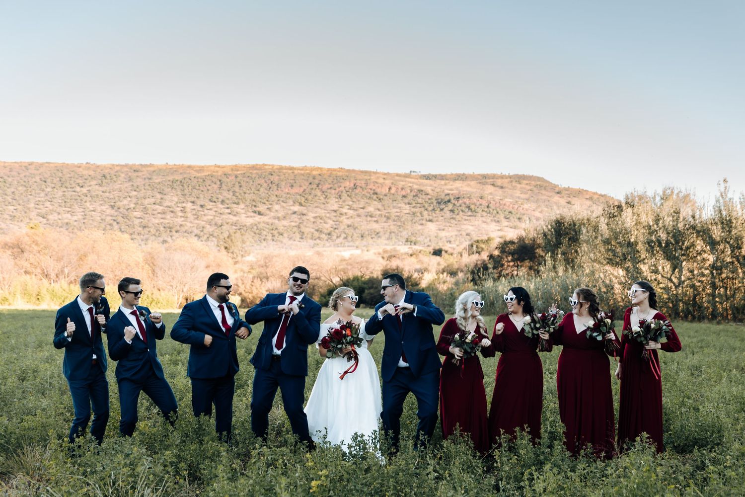 Wedding party in navy suits and burgundy dresses celebrate outdoors in a green field with rolling hills in the background.
