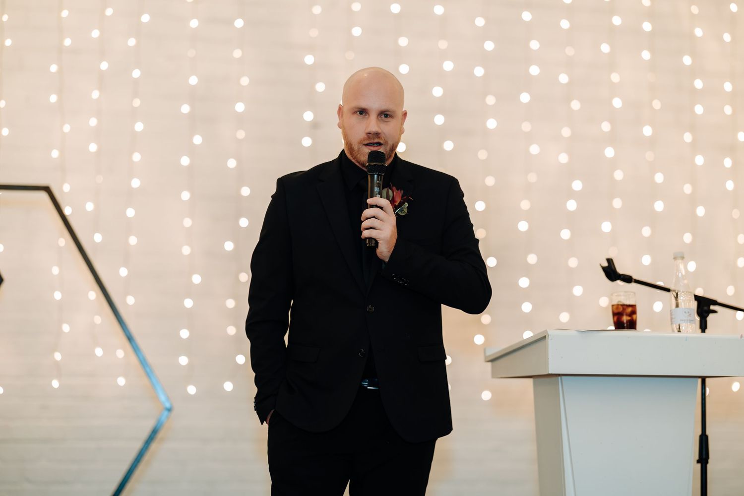 Bald man in black suit speaking into microphone at elegant event with bokeh fairy light backdrop.