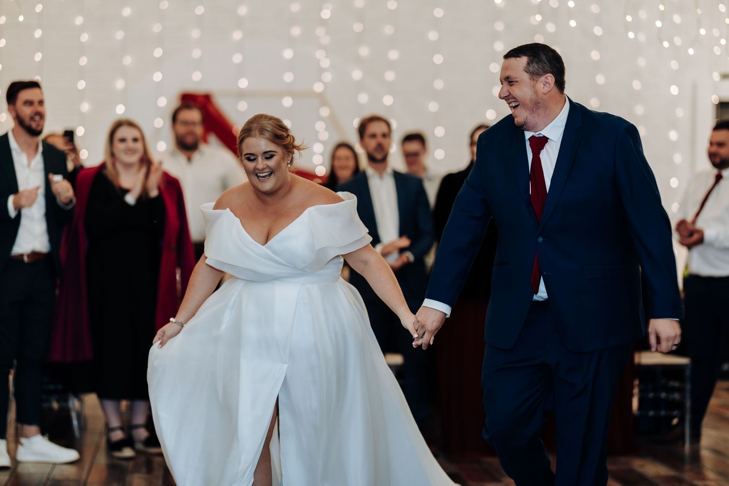 Bride and groom hold hands smiling during first dance at elegant wedding reception with fairy lights.