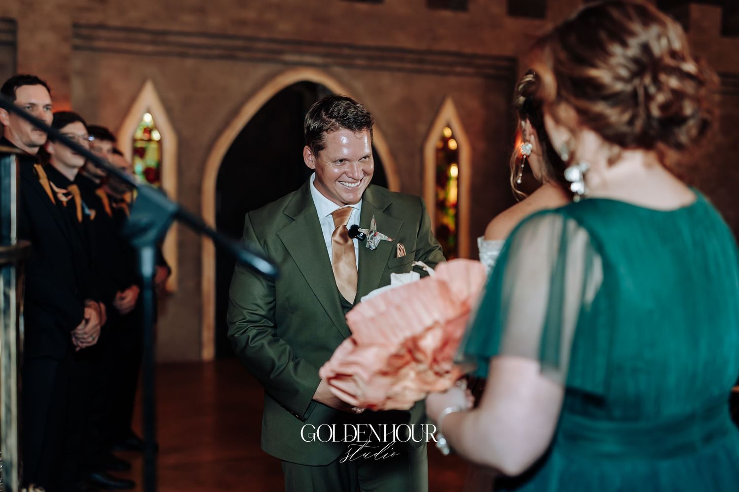 Smiling groom in green suit greeting guest holding pink bouquet at elegant wedding ceremony.