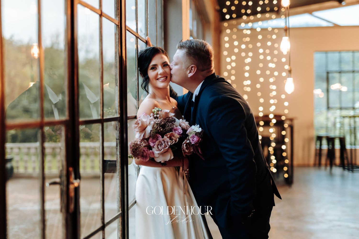 Groom in navy suit kissing bride on cheek as she holds a lush pink bouquet near large glass doors.