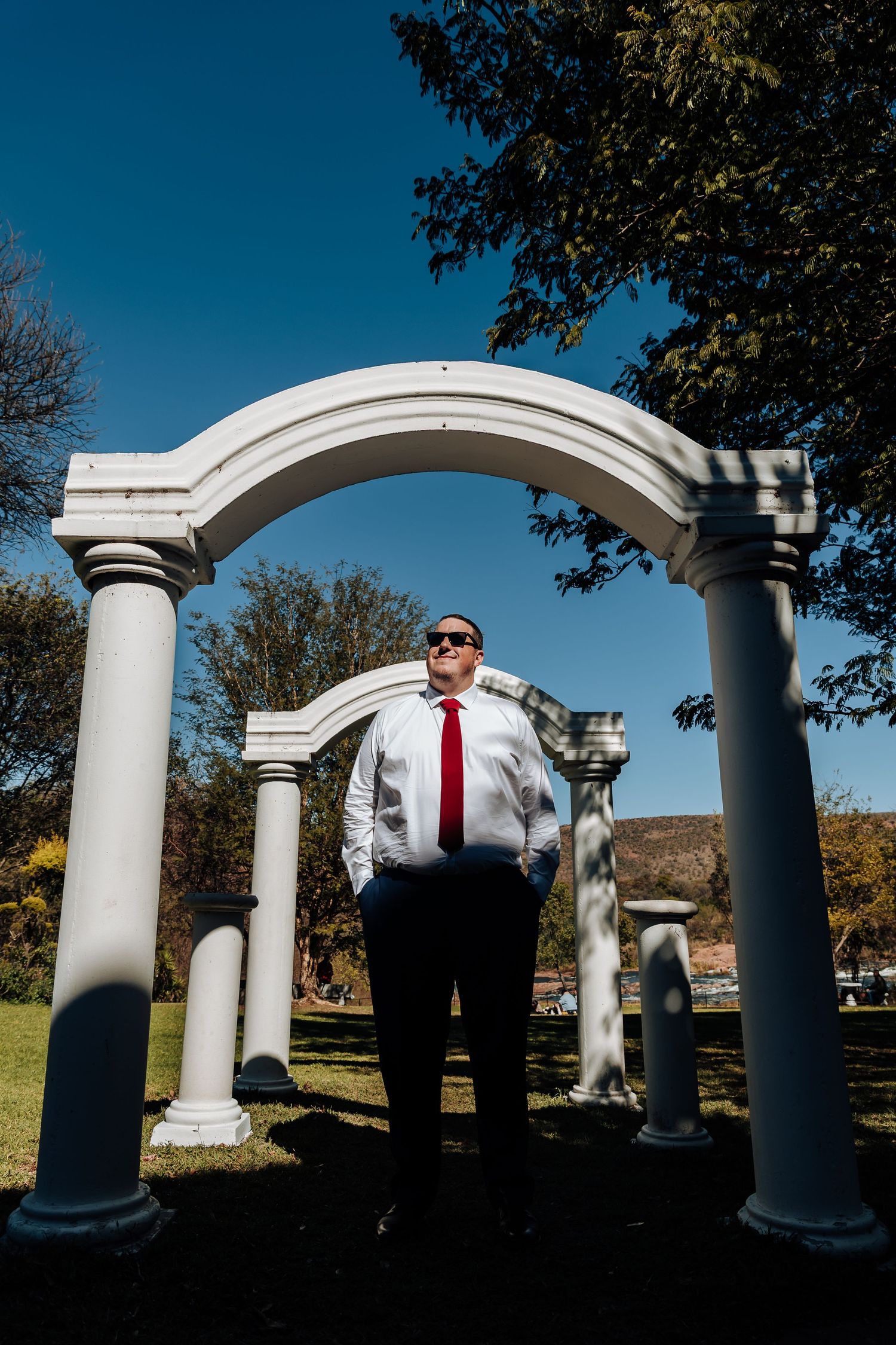 Man in white shirt and red tie standing under white arch columns in a sunny park with blue sky.