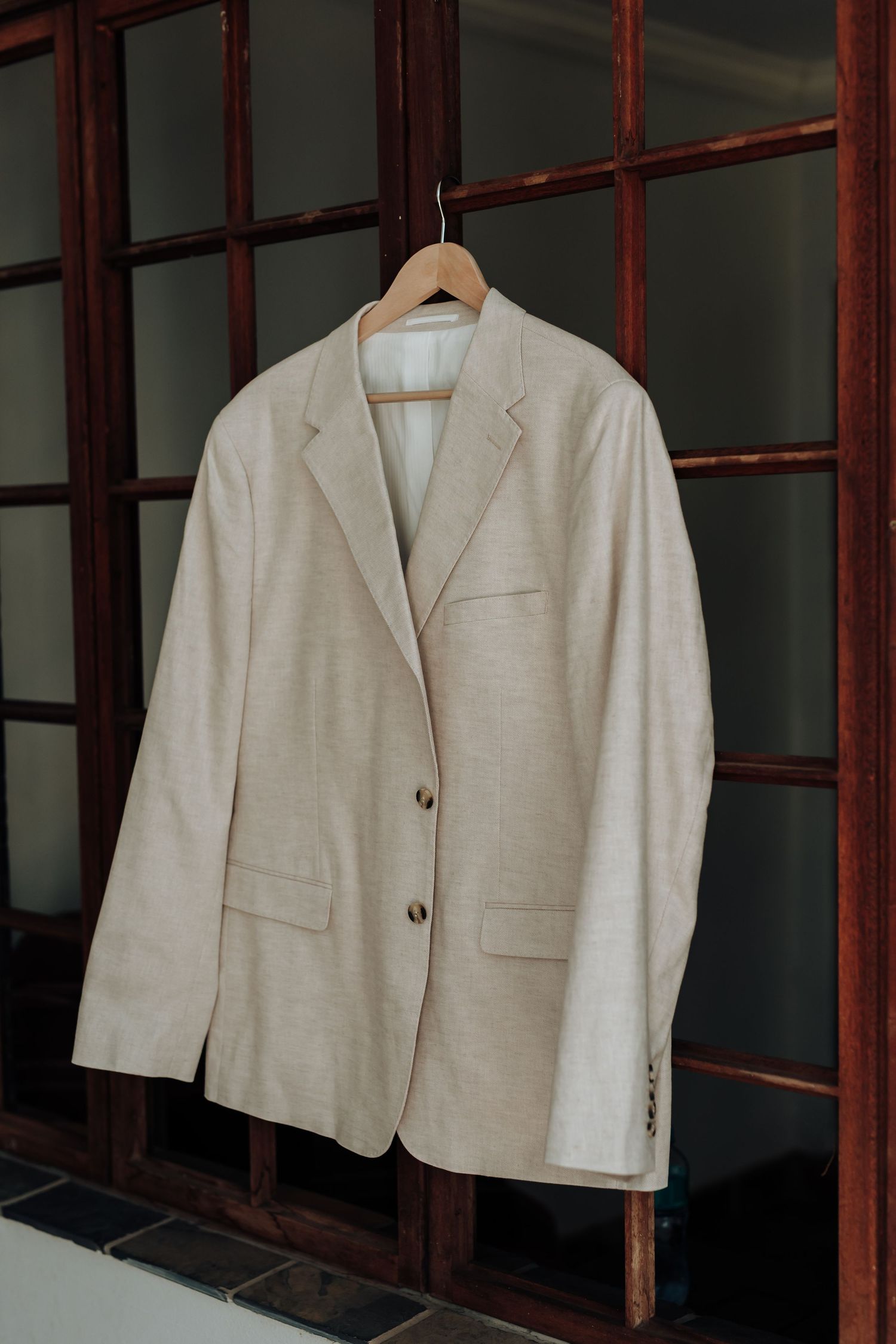 A cream-colored linen blazer hanging on a wooden hanger against a dark wooden wall panel.
