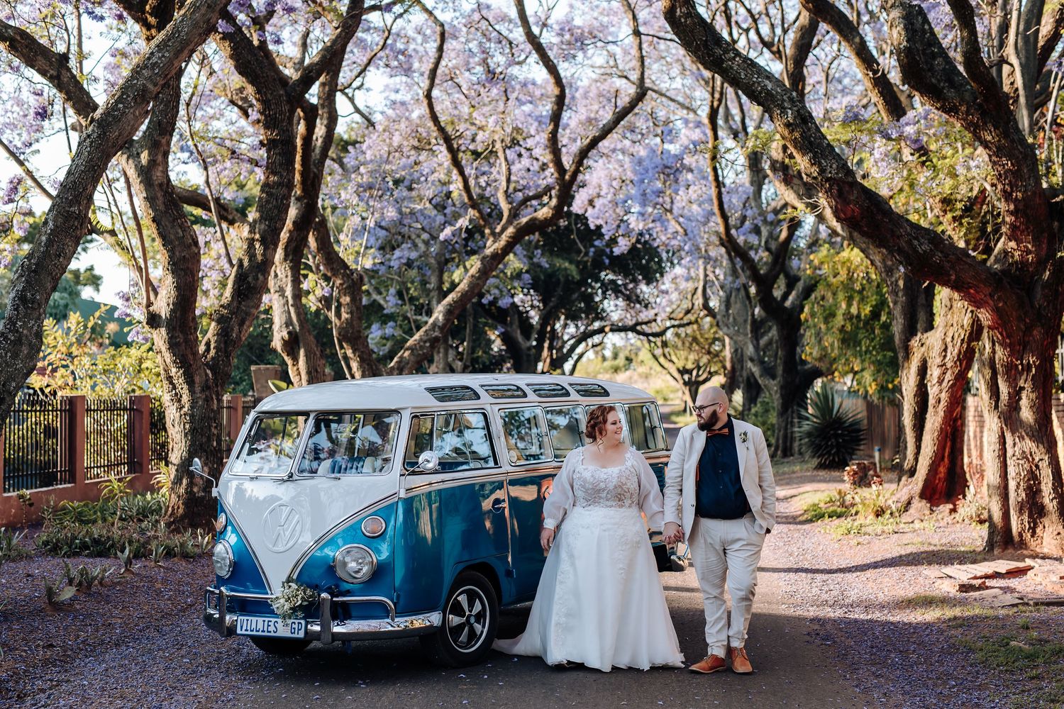 A vintage blue Volkswagen van parked under blooming trees on a scenic path during a wedding celebration.