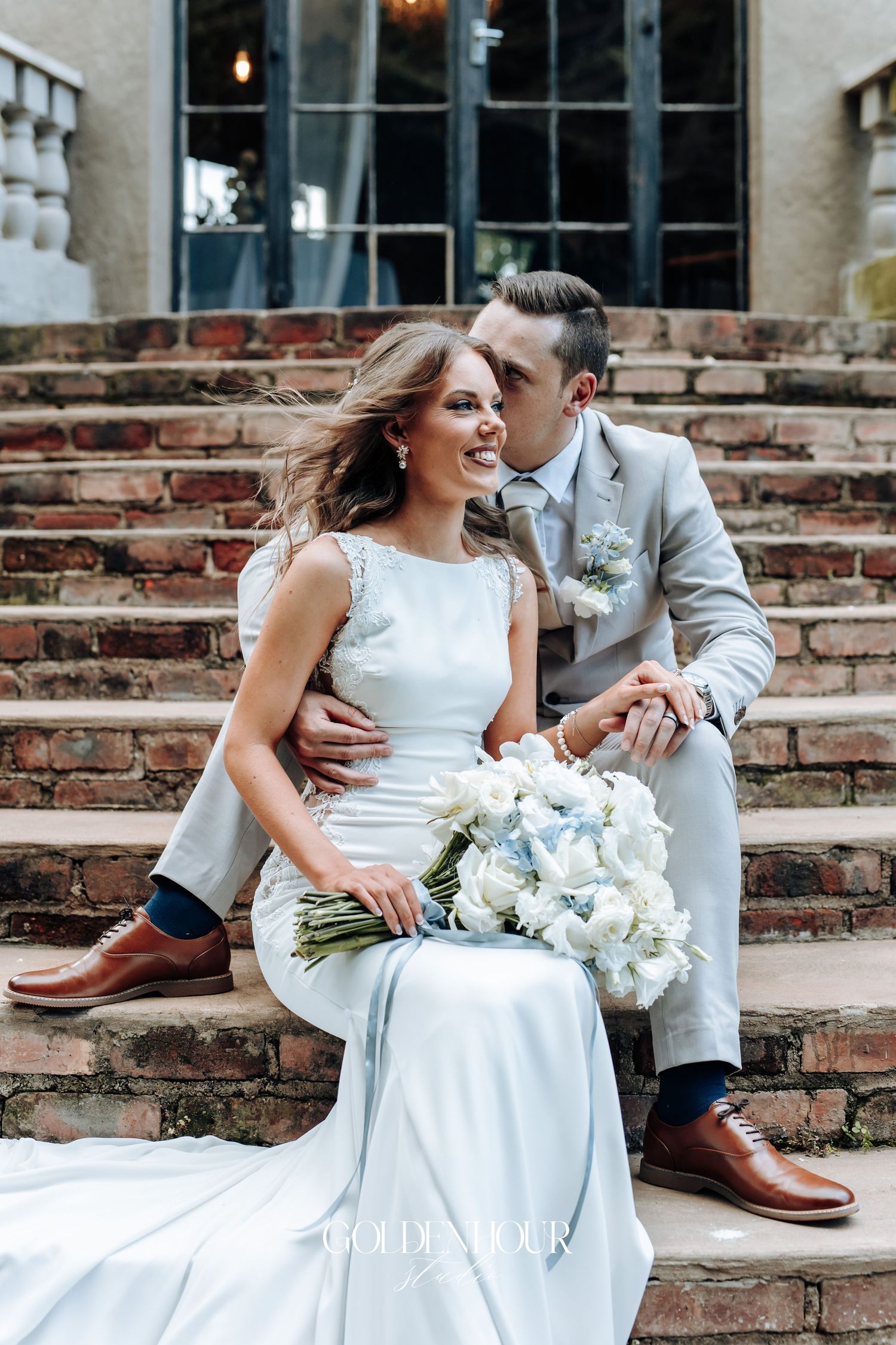 Wedding couple sitting on brick steps with white bouquet and elegant attire.
