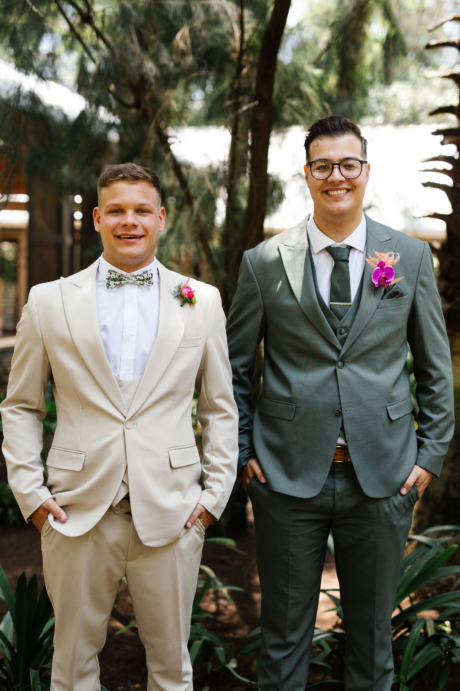 Two people in contrasting suits stand together outdoors among tropical foliage and trees.