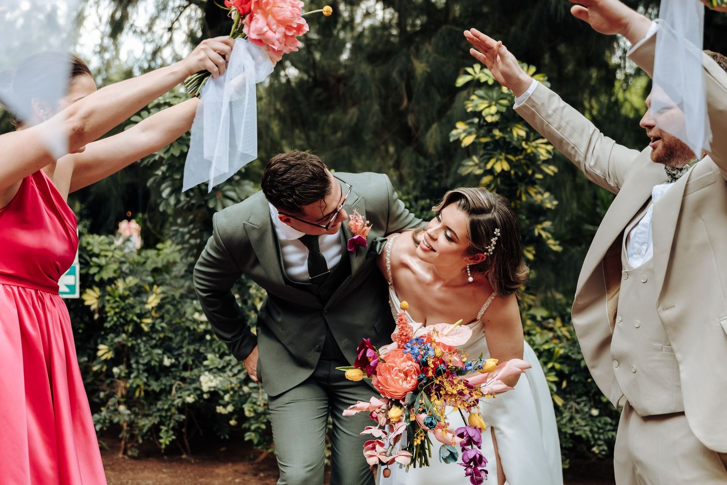 Wedding party members dance and celebrate on a garden path surrounded by greenery.