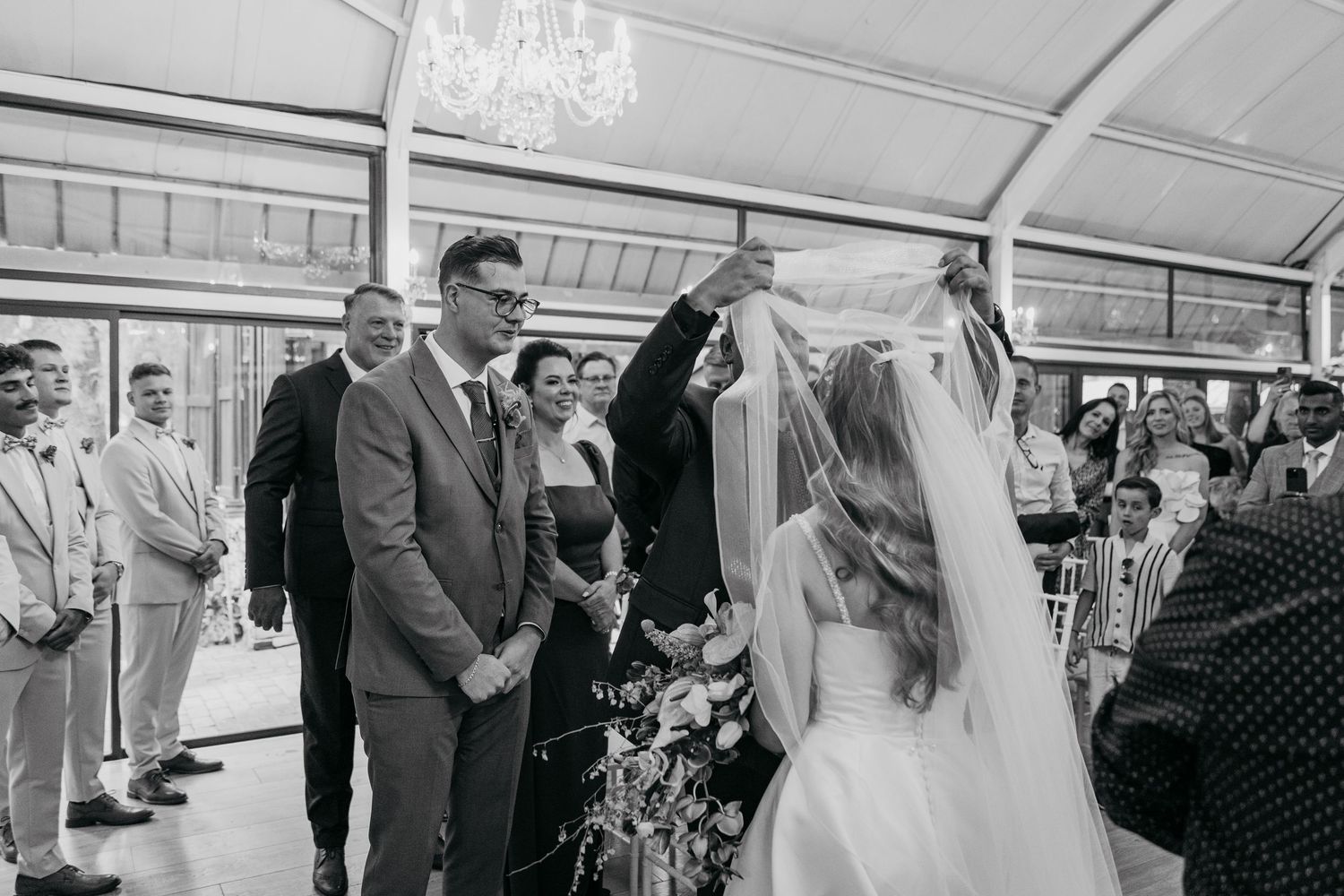 A black and white wedding ceremony photo showing guests gathered under a covered outdoor venue.