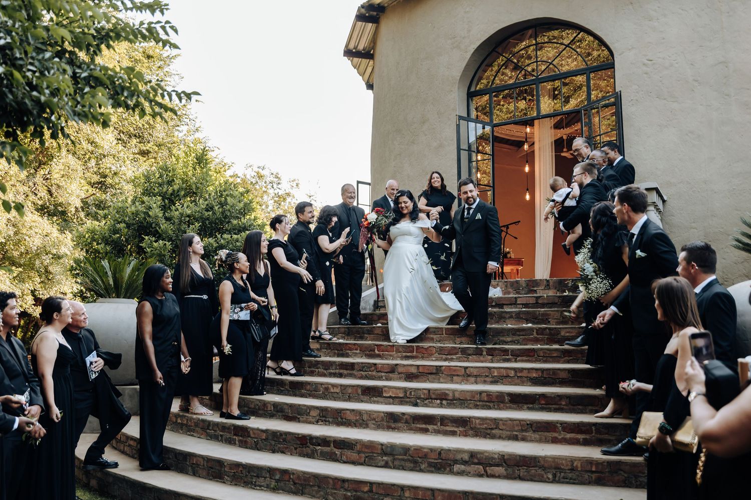 Wedding party stands in formal attire on stone steps leading to ornate church entrance during outdoor ceremony.