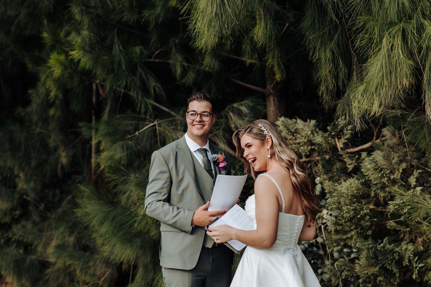 A romantic couple embraces outdoors surrounded by lush evergreen trees in a forest setting.