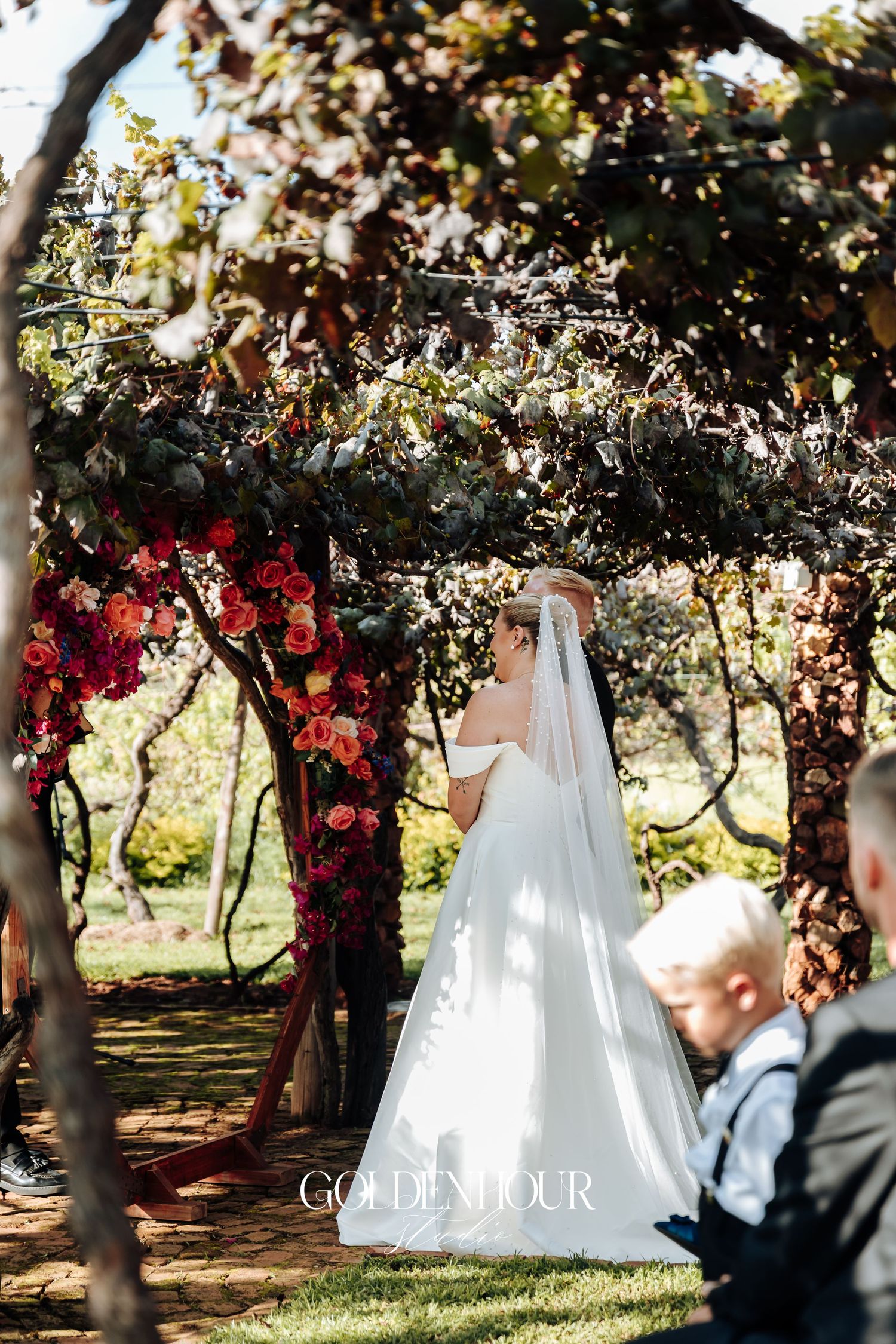 Romantic outdoor wedding ceremony under a floral-covered pergola with red roses and greenery in a garden setting.