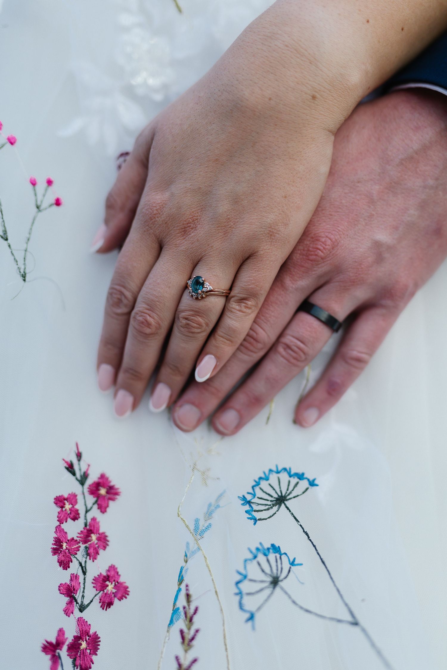 Hands wearing wedding rings rest on a white surface decorated with delicate pink and blue floral artwork.