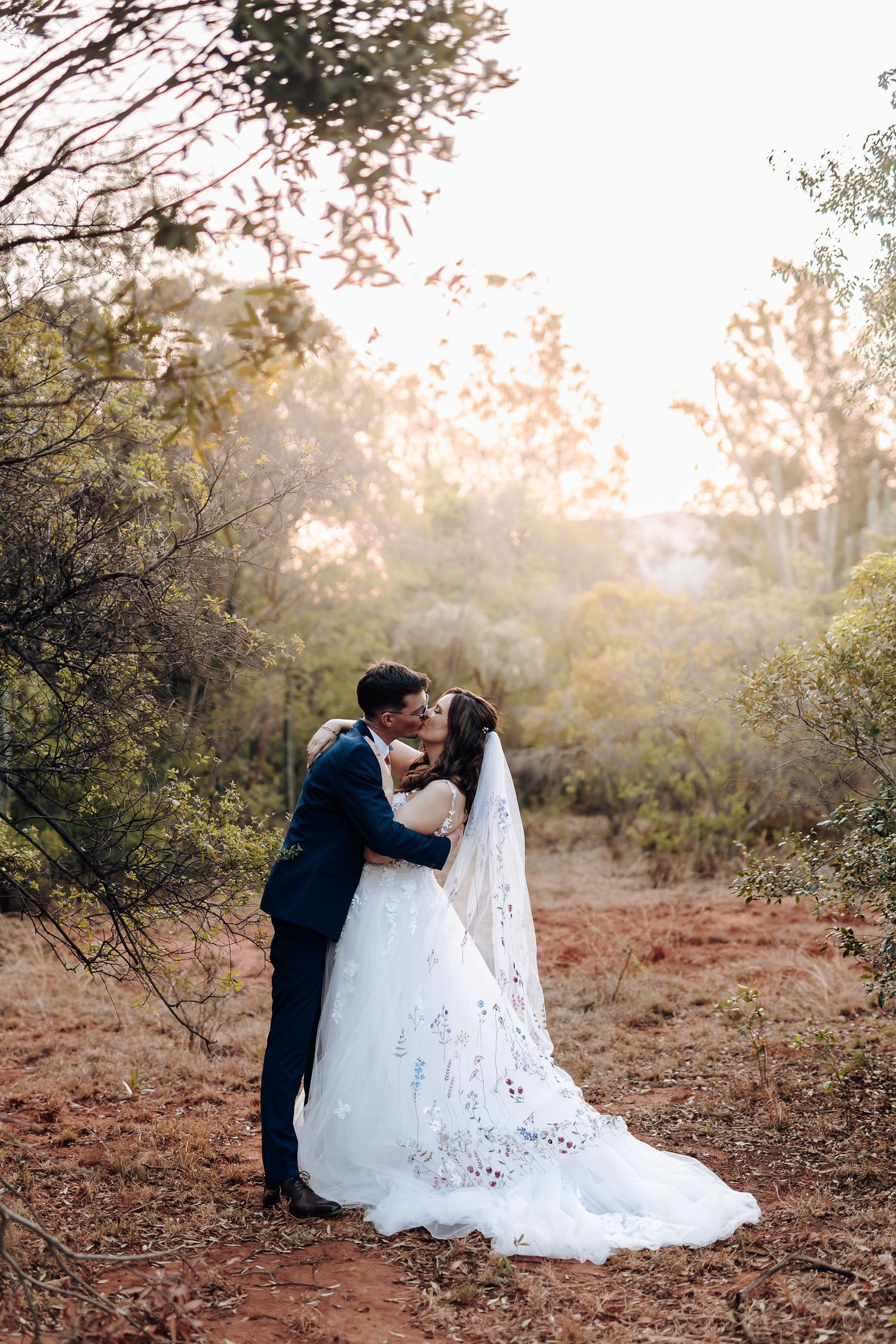 A romantic wedding couple shares a tender moment in a sunlit outdoor setting with autumn foliage.