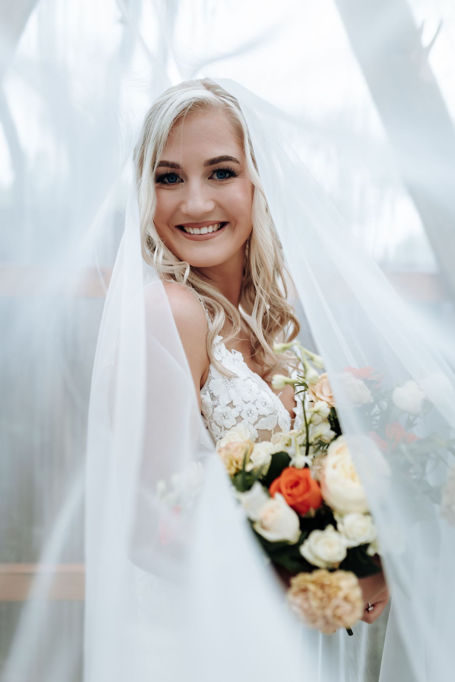 A bride in a white wedding dress and veil holds a colorful bouquet of roses and hydrangeas against a soft, misty background.