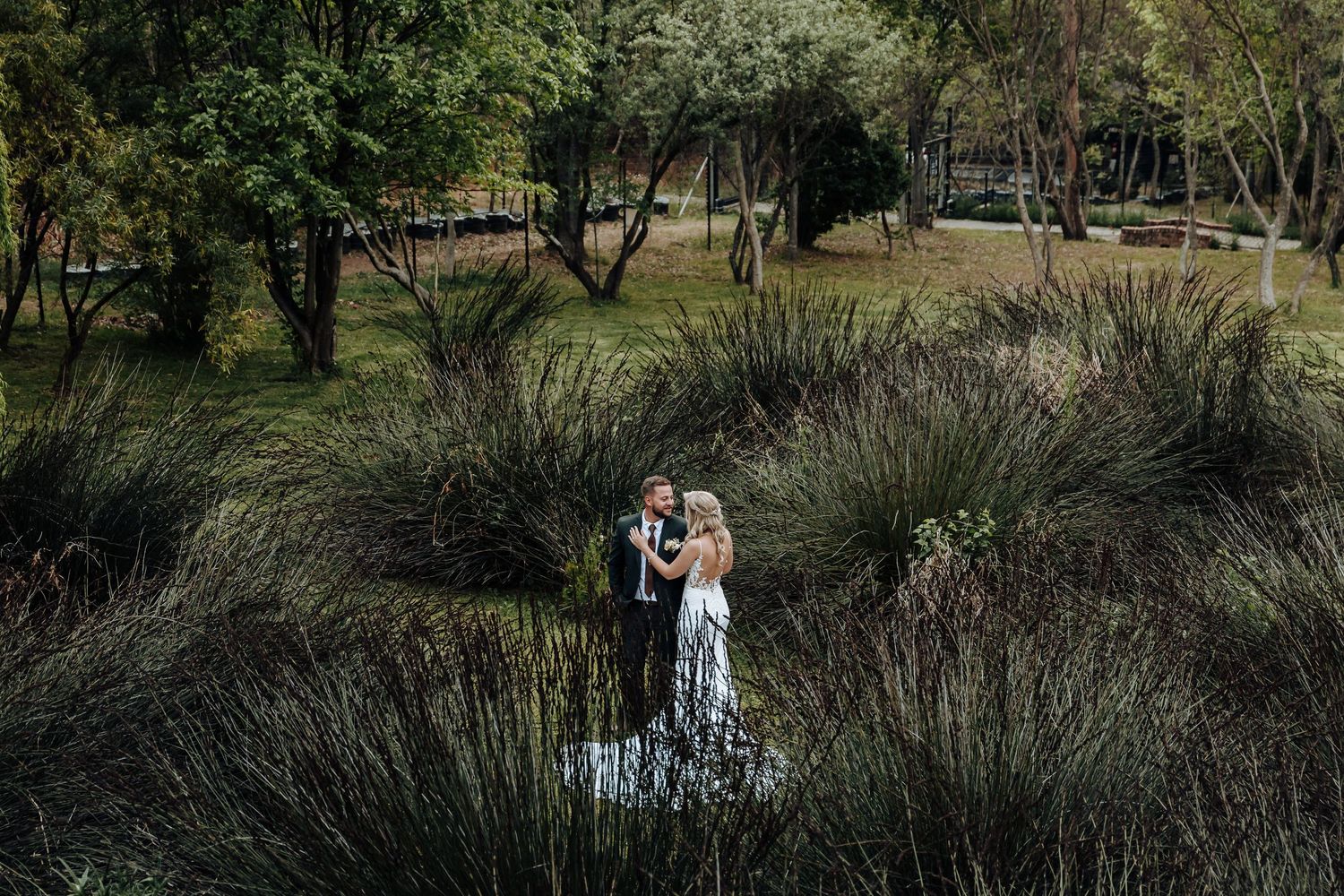 Couple in formal wedding attire embracing in a lush garden setting with natural greenery and trees in the background.
