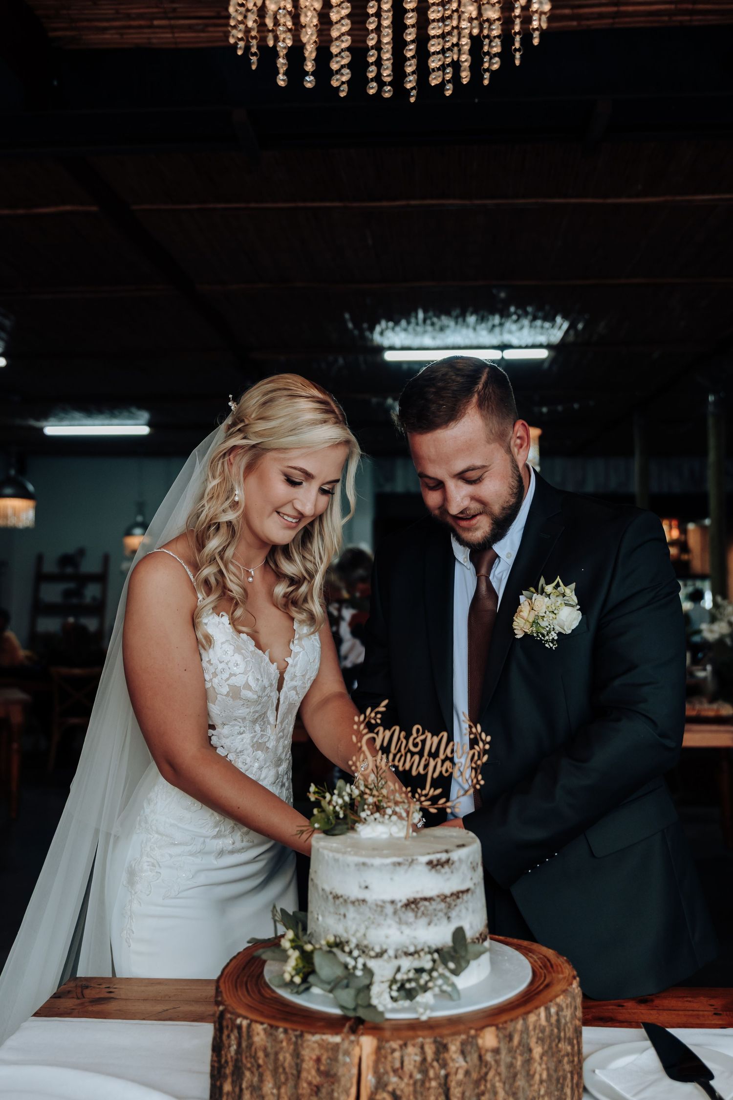 A couple cutting their elegant white wedding cake together at their reception under a crystal chandelier.