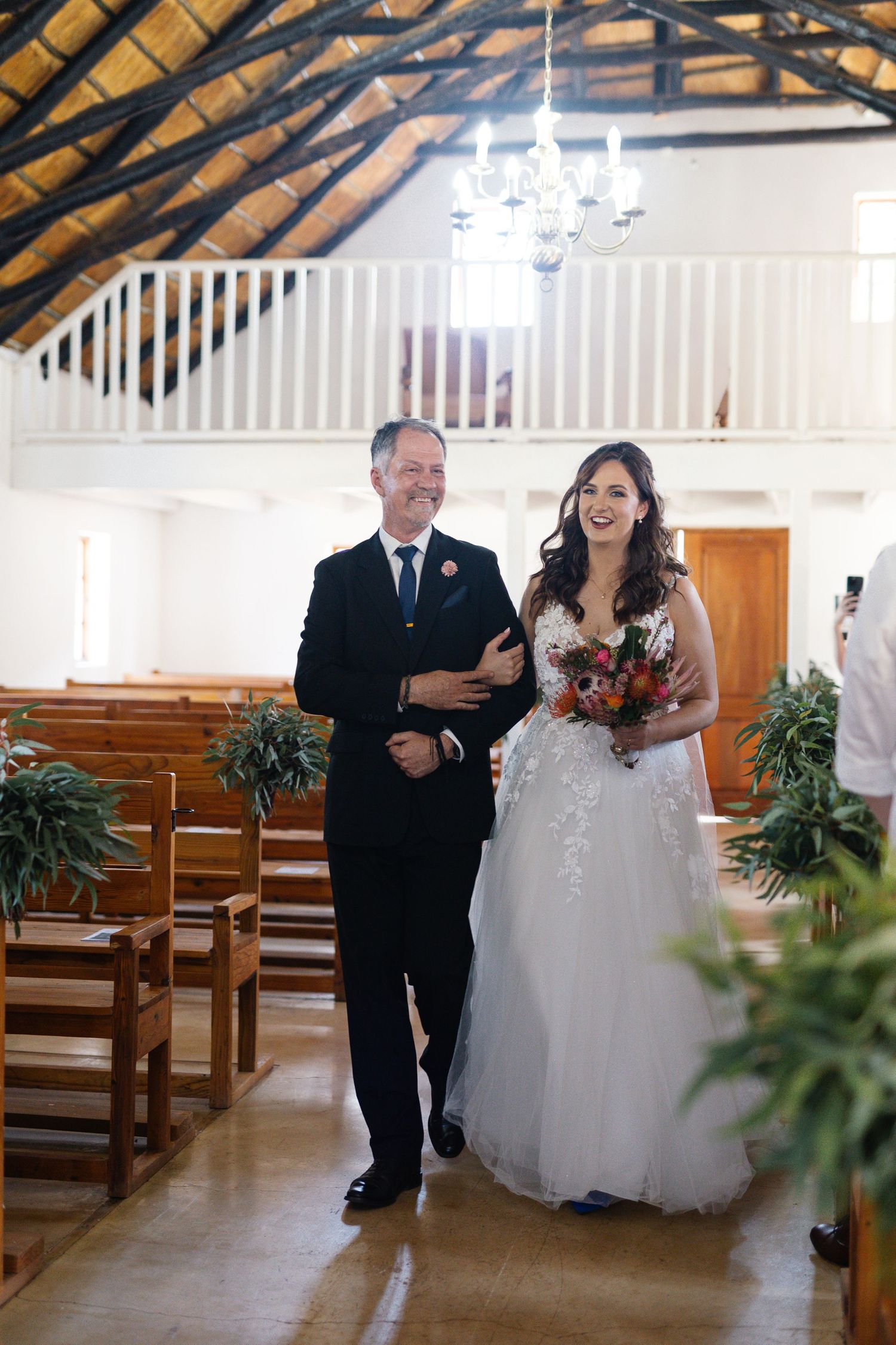 A joyful wedding party walks down the aisle of a bright chapel with wooden beams and pews.