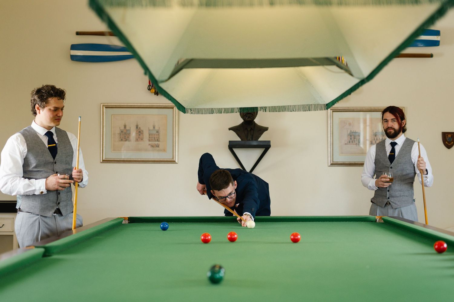 Three people in formal attire playing snooker on a green table in a classic game room with framed artwork on the wall.
