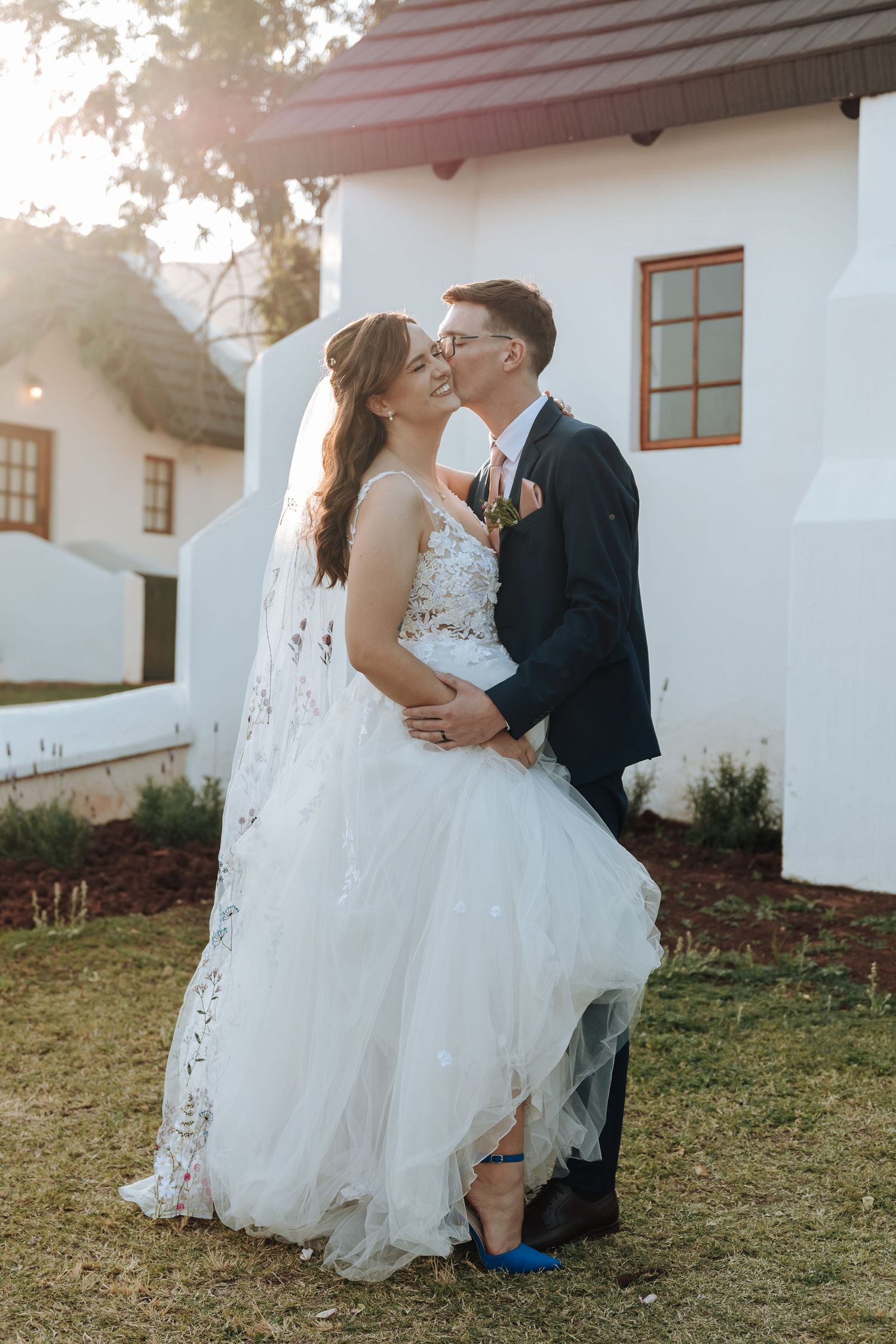 A romantic wedding couple shares tender moments in a garden setting with white buildings in the background during sunset.