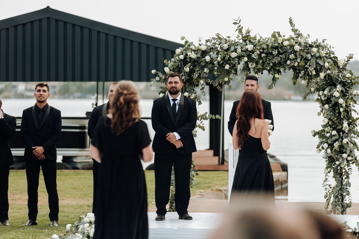 Wedding ceremony takes place under a floral arch by a serene lake with guests gathered to witness the occasion.