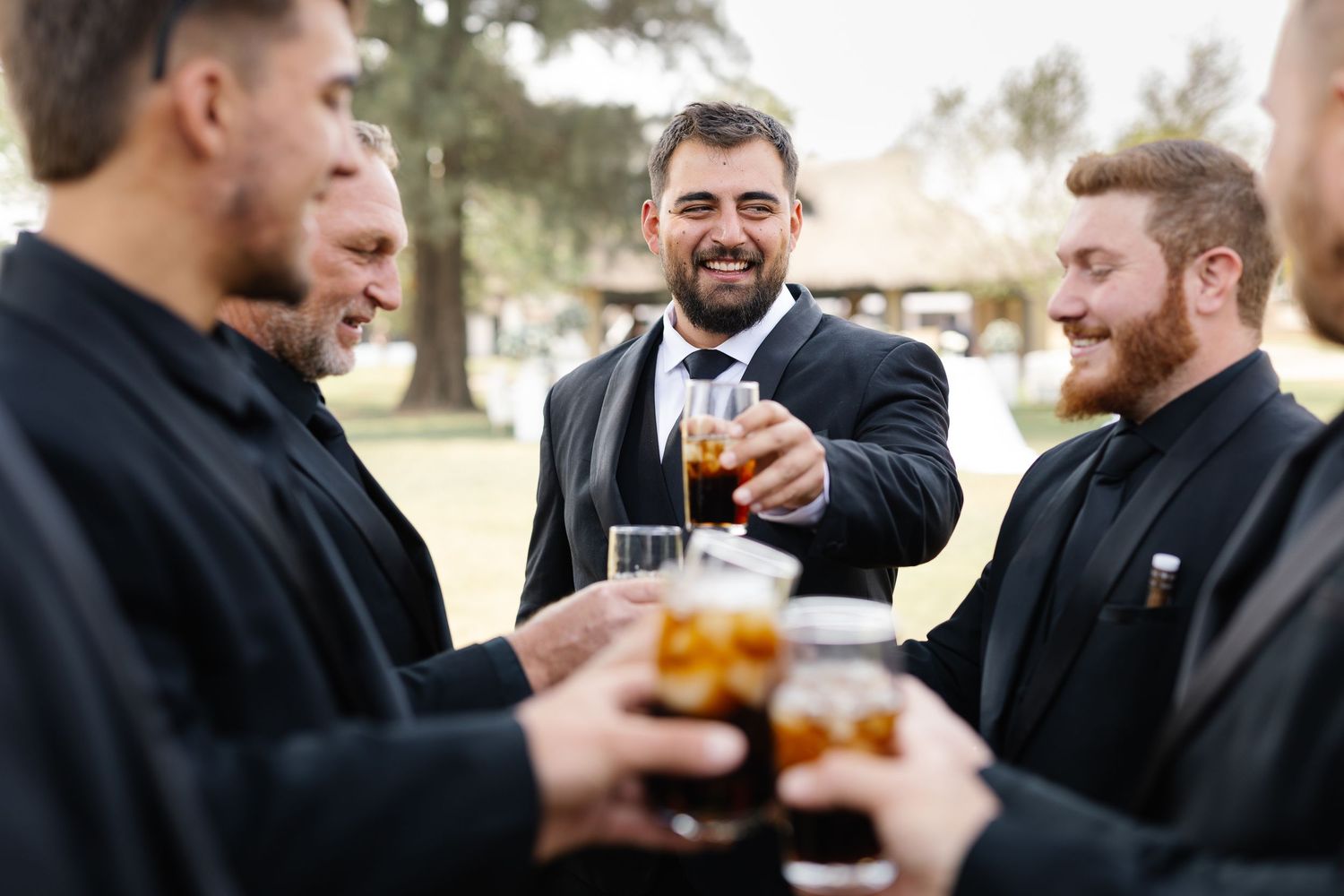 A group of men in dark suits toast with whiskey glasses while celebrating outdoors.