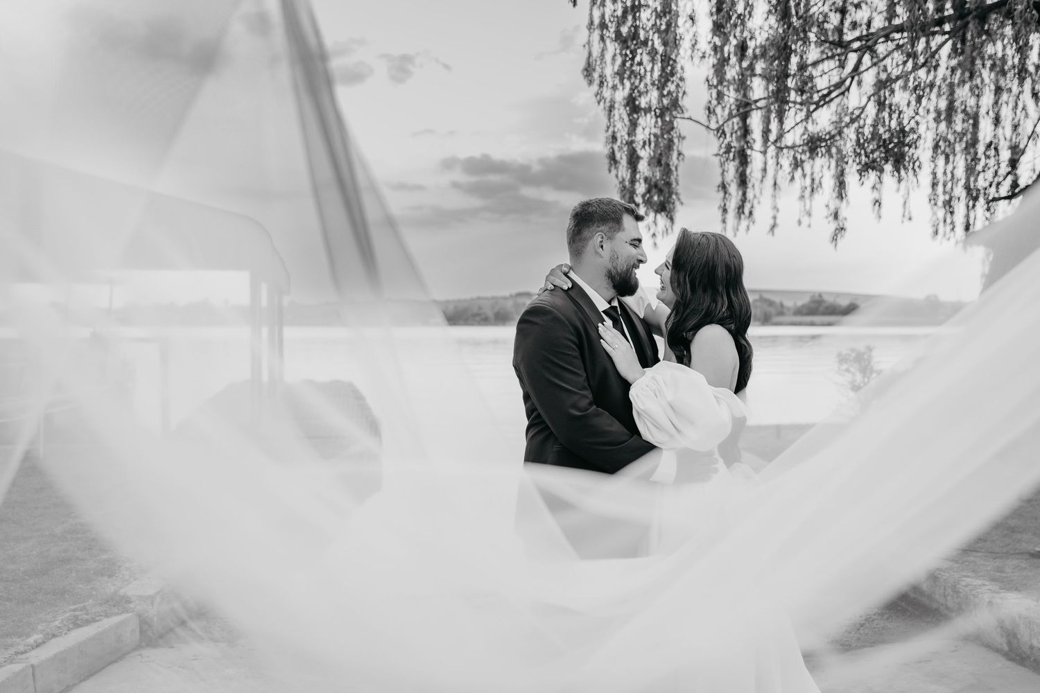 A romantic couple embraces under a willow tree during a dreamy outdoor wedding photoshoot.