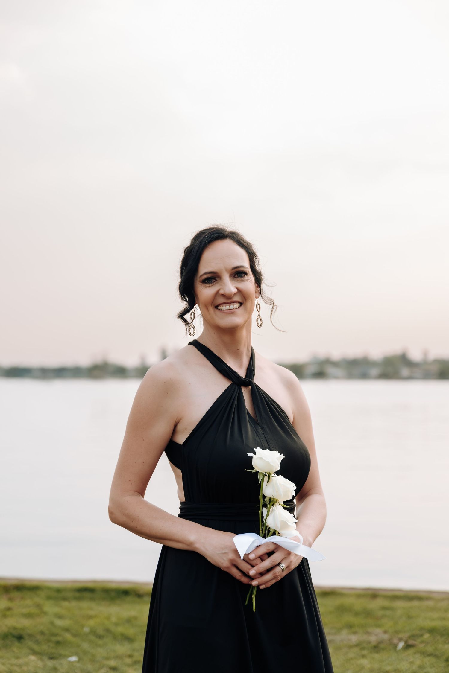 Person in black halter dress smiles while holding white roses near a lake during golden hour.