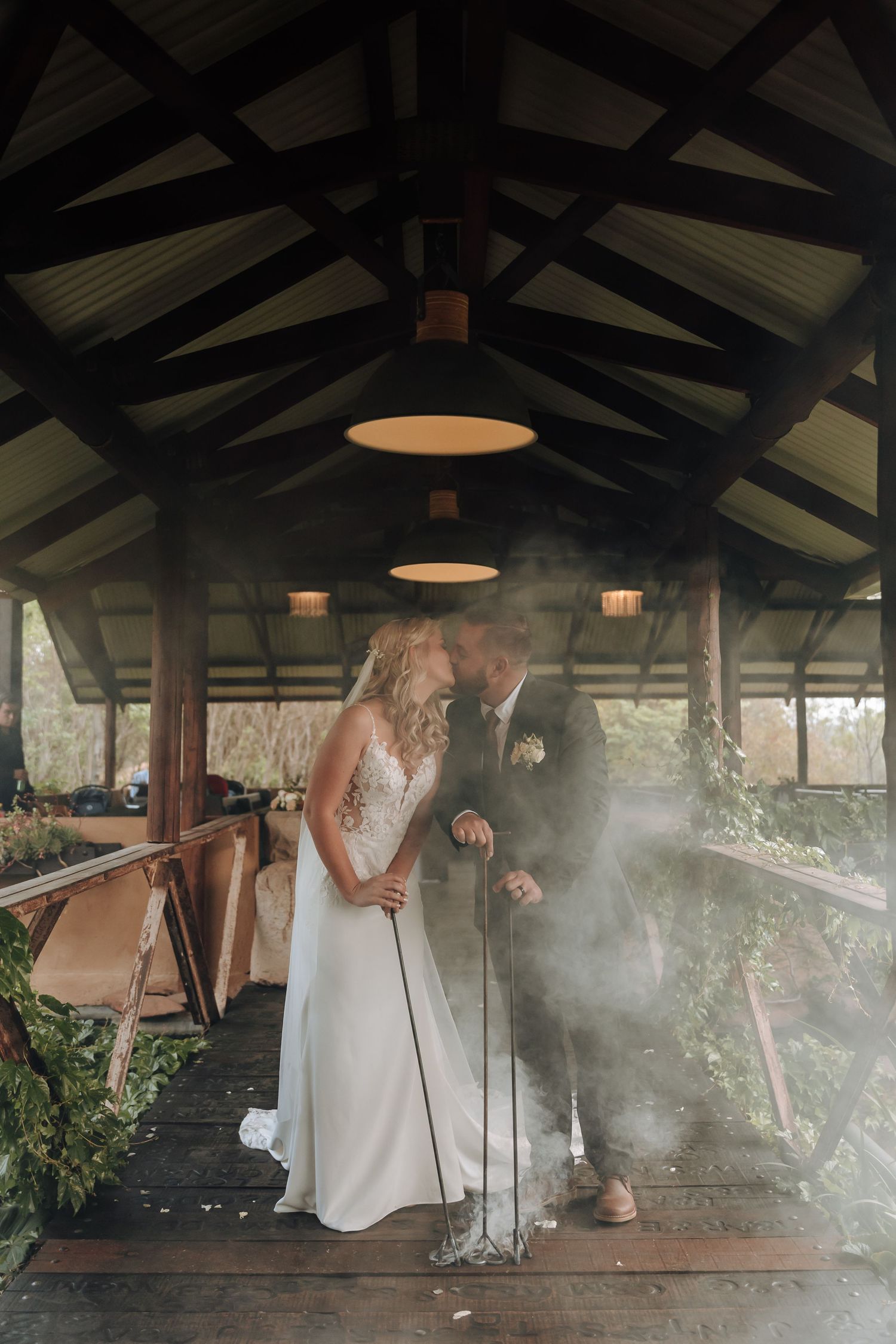 A romantic wedding moment captured under a rustic wooden pavilion with soft lighting and atmospheric mist.