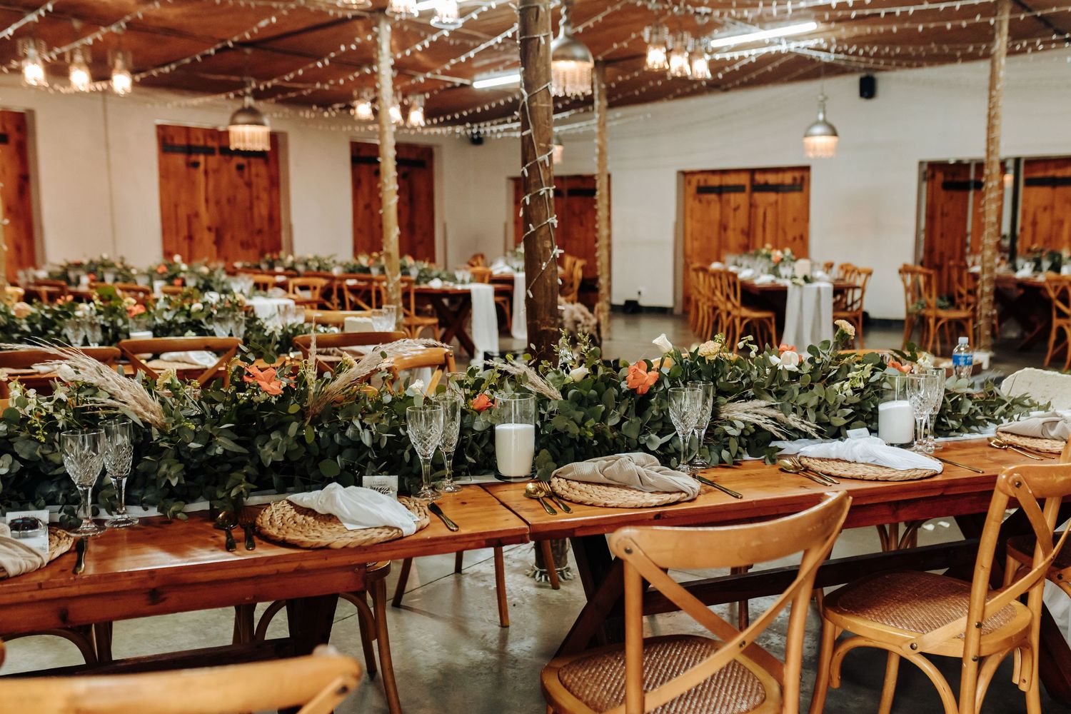 Long wooden banquet tables adorned with lush greenery and candlelight create an elegant rustic wedding reception setting.
