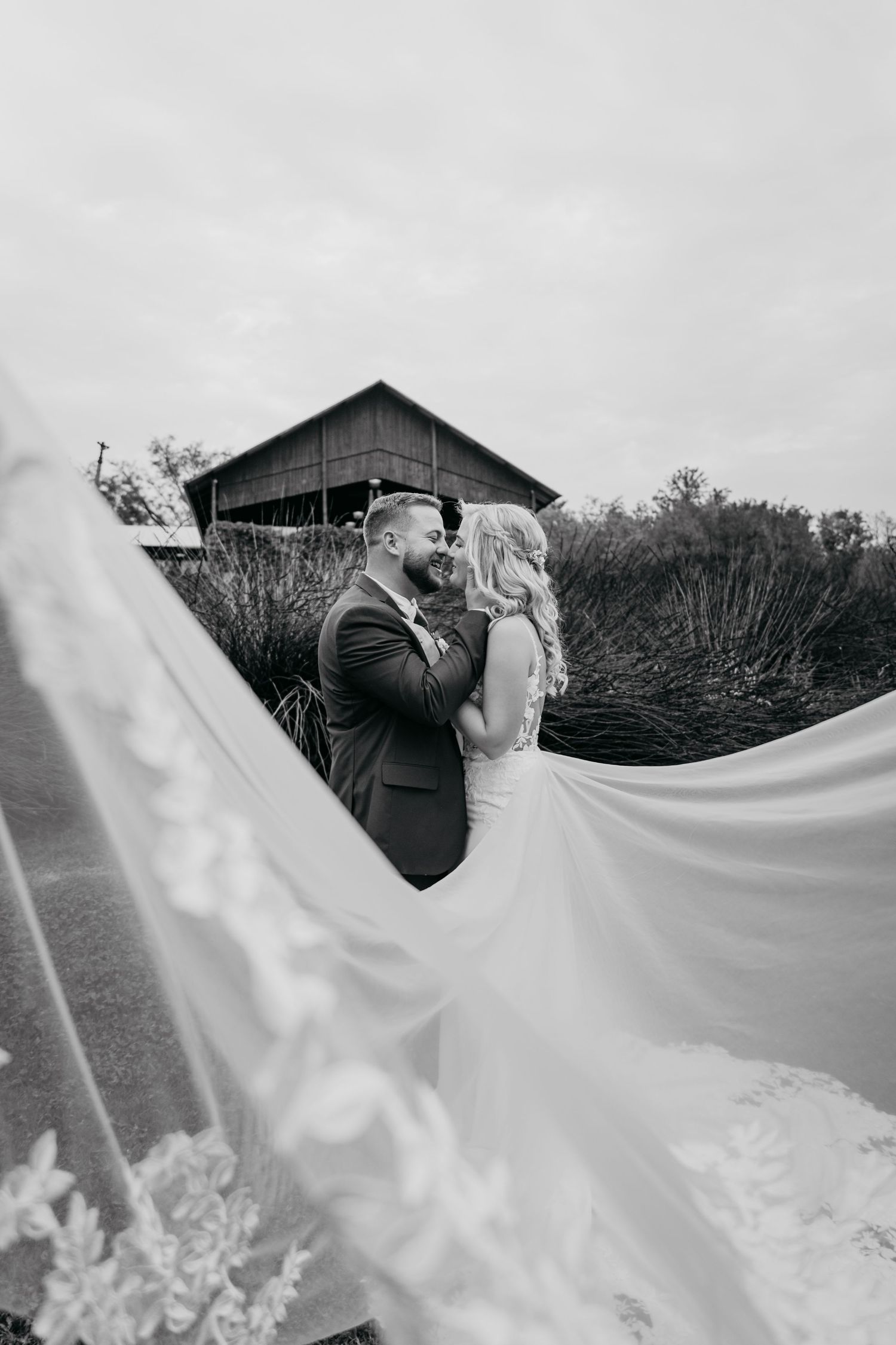 Black and white wedding photo with flowing white dress veil in foreground and rustic barn building in background.