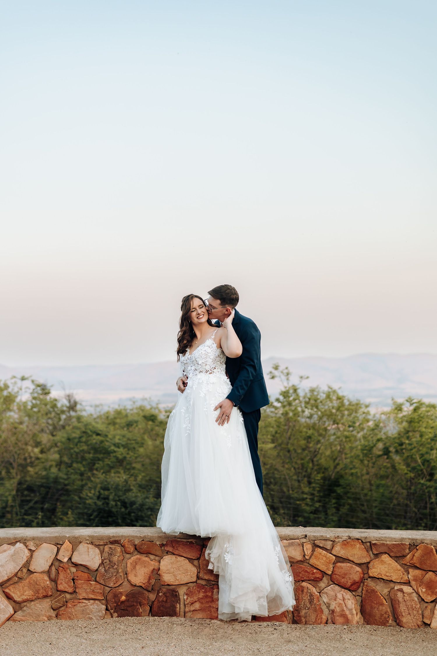 Romantic couple in wedding attire embraces against a stone wall with trees and sunset sky in the background.