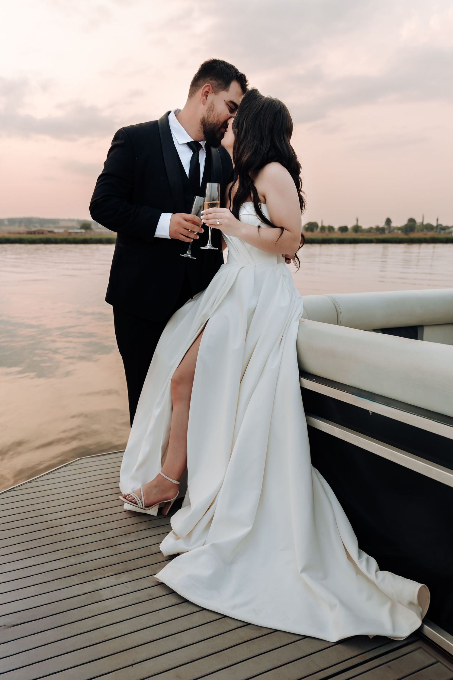 Newlywed couple sharing champagne toast on boat deck at sunset with elegant white wedding dress and black suit.