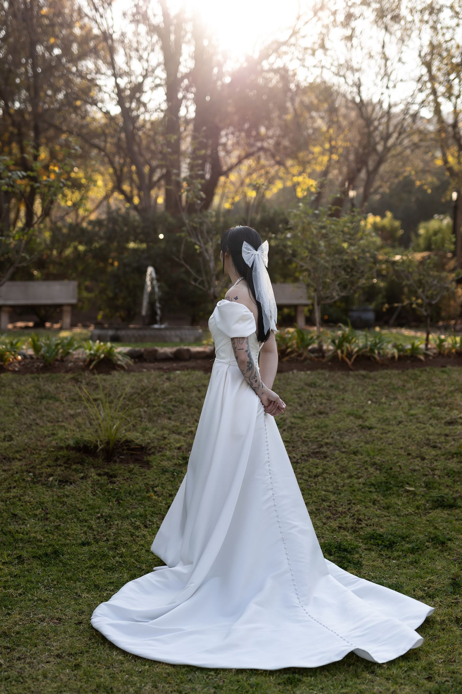 A white wedding gown with a flowing train is showcased in a sunlit garden setting at dusk.