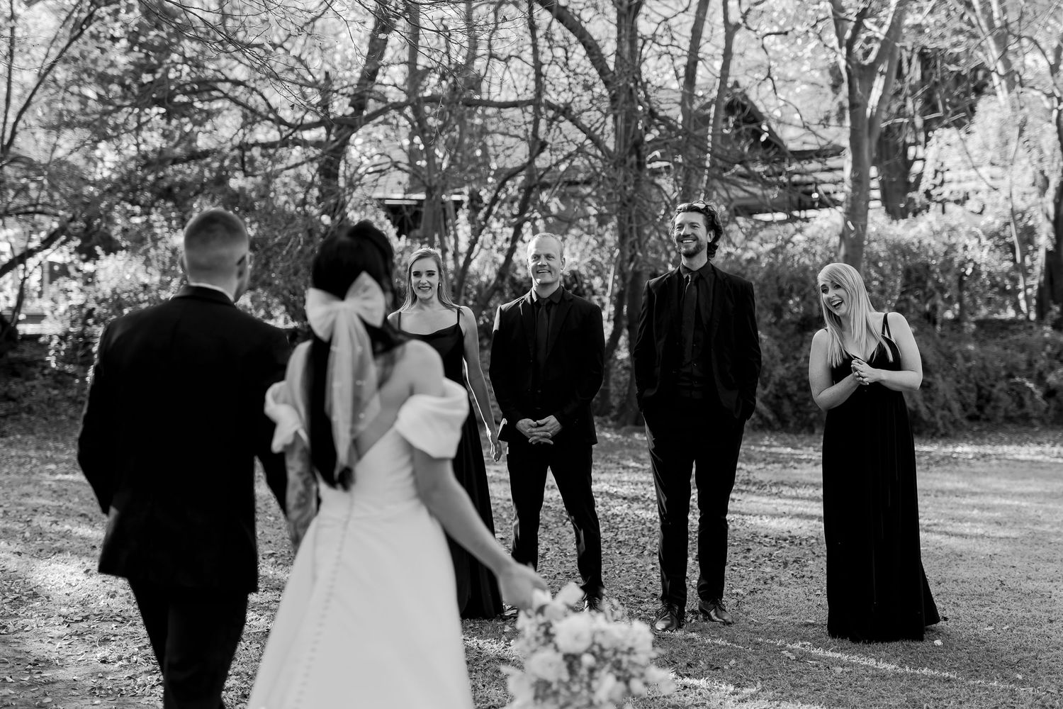A wedding party gathered together in a black and white photo, with trees and natural surroundings in the background.